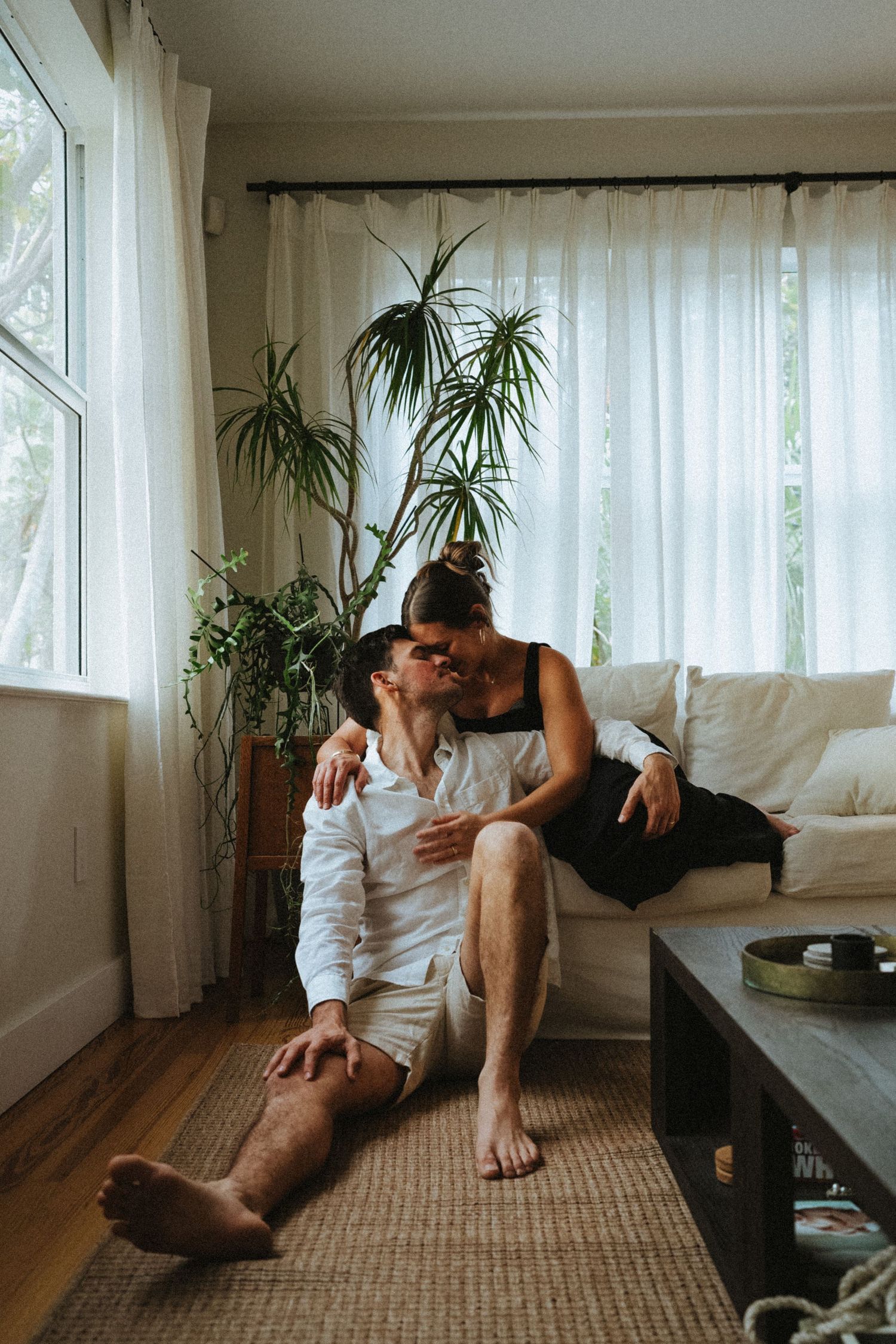 A couple shares a tender kiss in a cozy living room with white curtains and lush green plants.