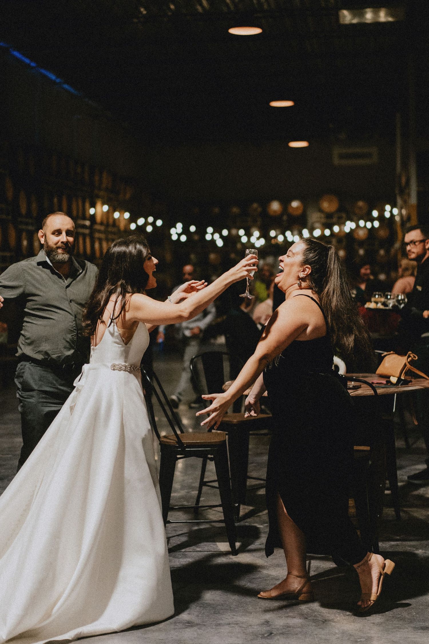 Bride toasting drinks with a guest in a black dress at an indoor wedding reception with string lights.