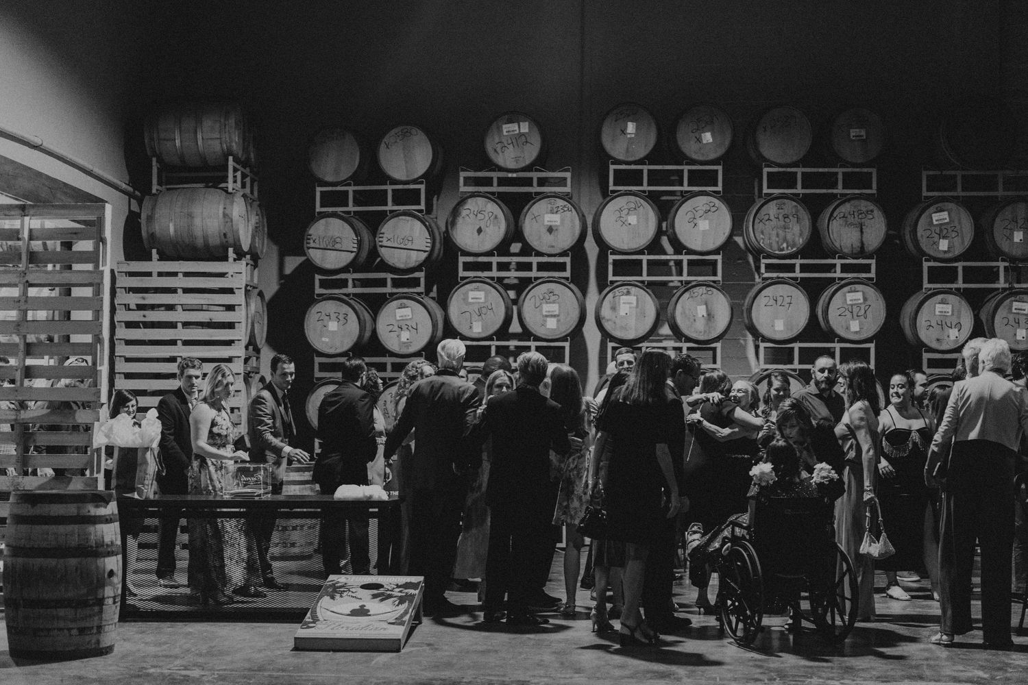 Guests mingle among wine barrels at a winery tasting event in a rustic barrel room.