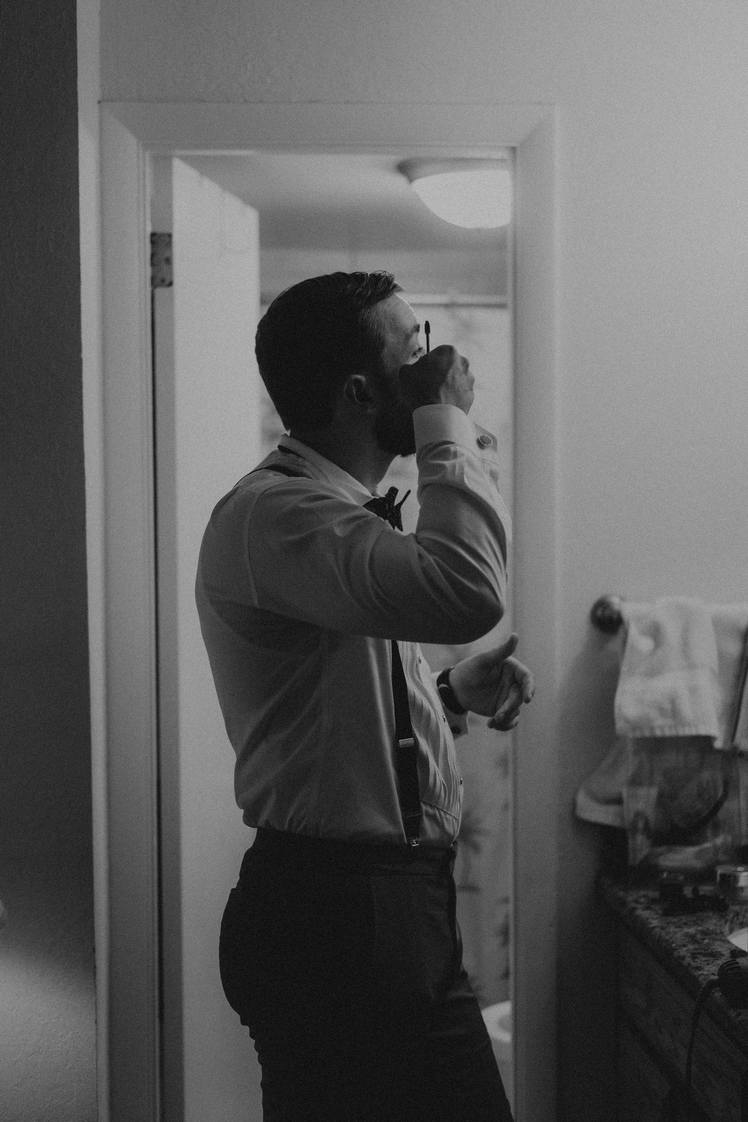 Groom adjusting bow tie in bathroom mirror while getting ready for wedding.