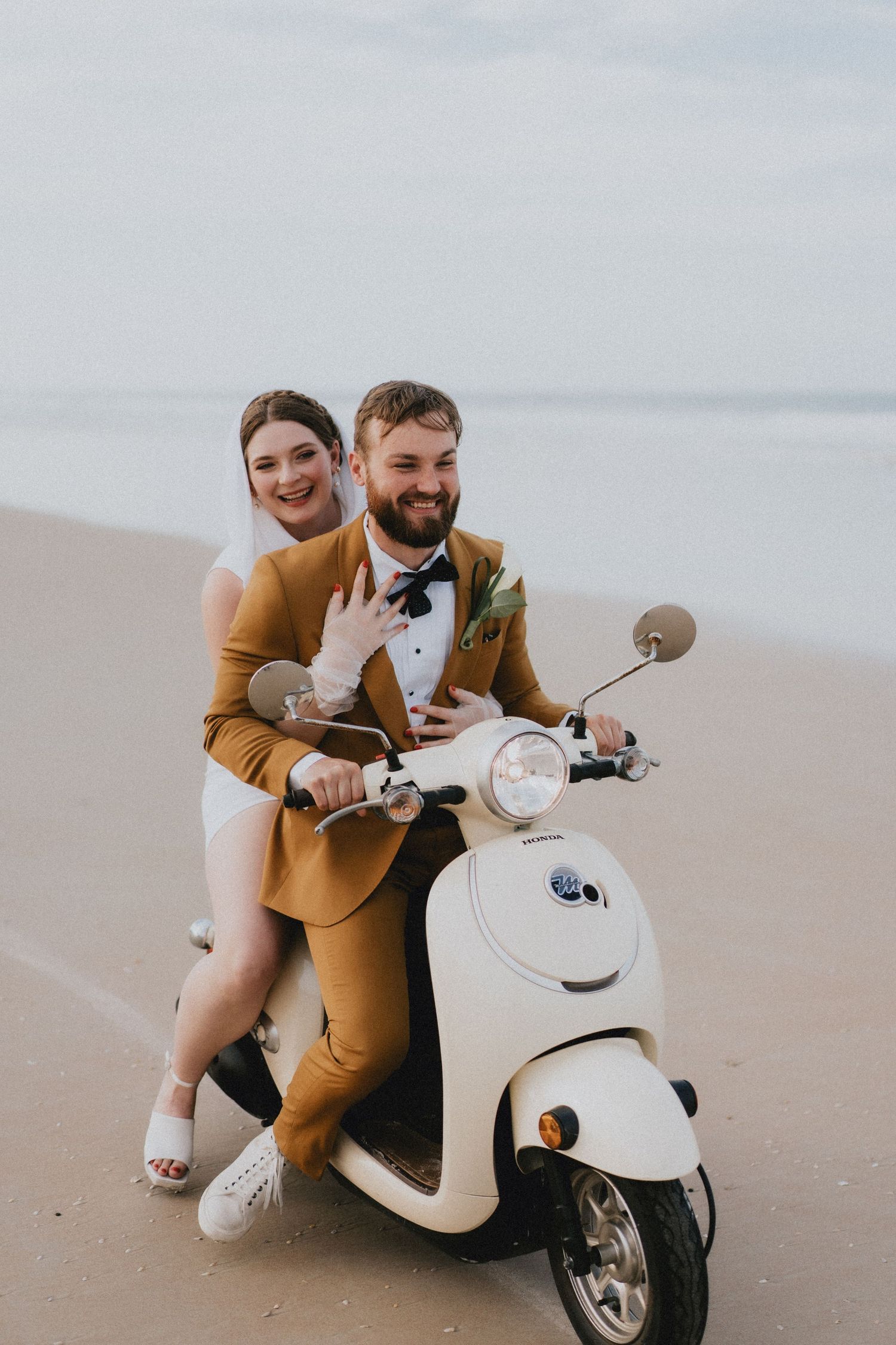 Happy bride and groom riding white vintage scooter together on sandy beach.