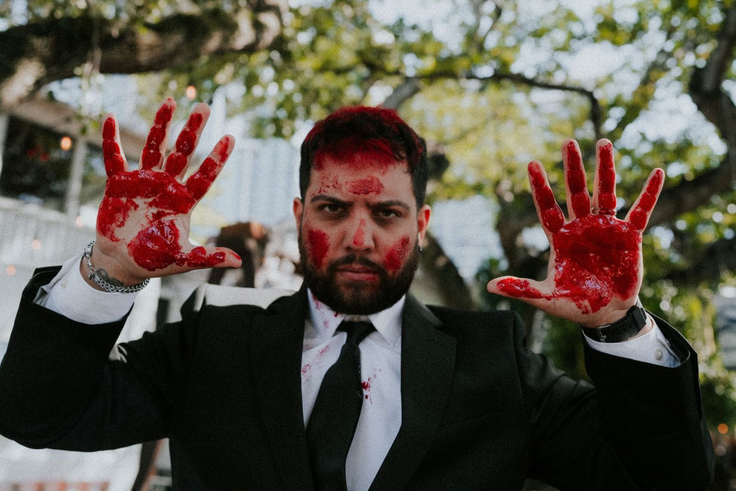 Man in black suit with red paint on face and hands raised, outdoor Halloween costume photo.