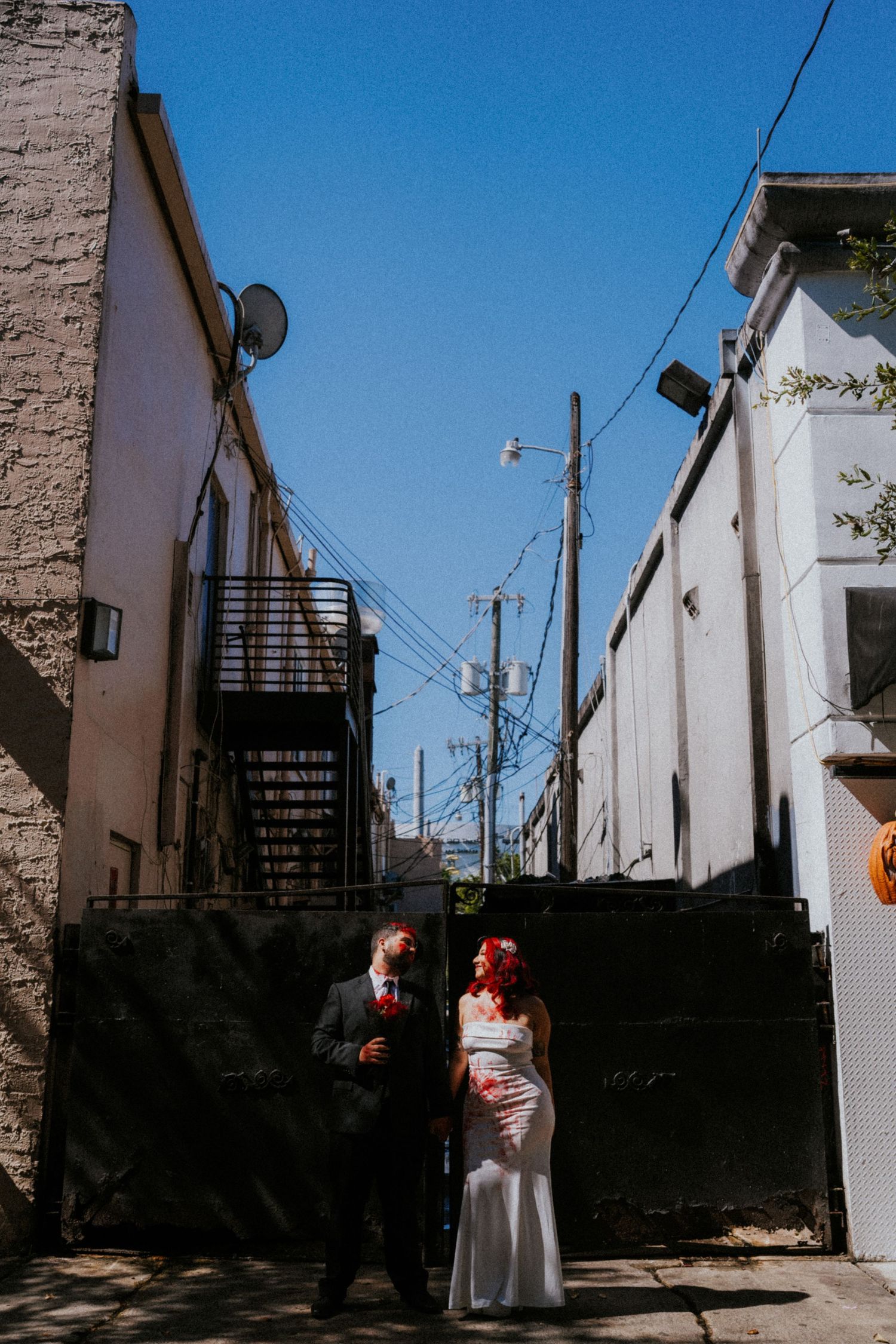 A bride and groom stand in an urban alley between buildings under a clear blue sky on their wedding day.