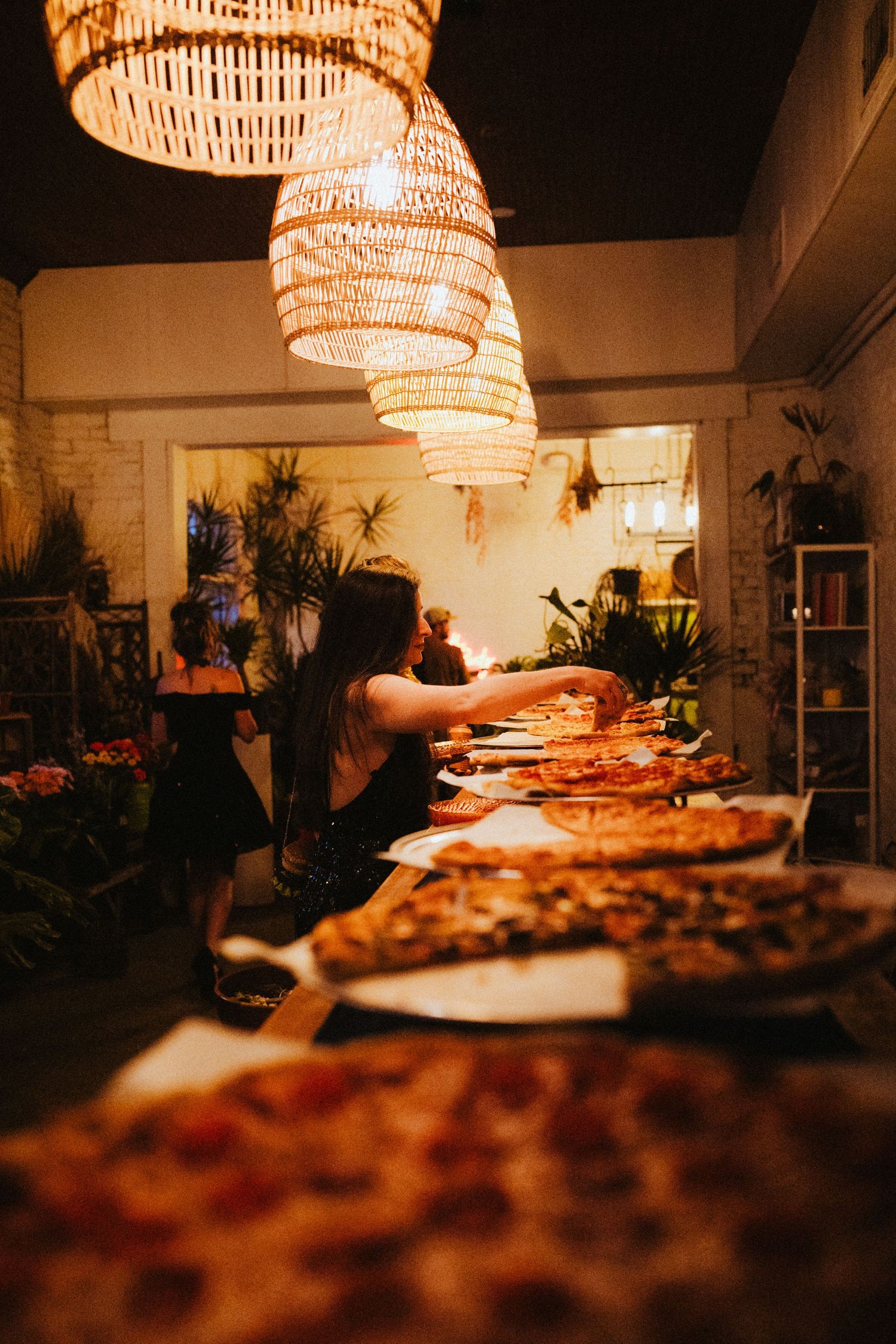Woman serving pizza at a long buffet table under warm wicker pendant lights in a stylish venue.