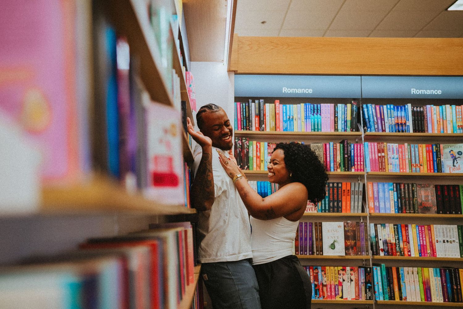 Couple laughing and flirting between colorful bookshelves in a cozy bookstore romance section.