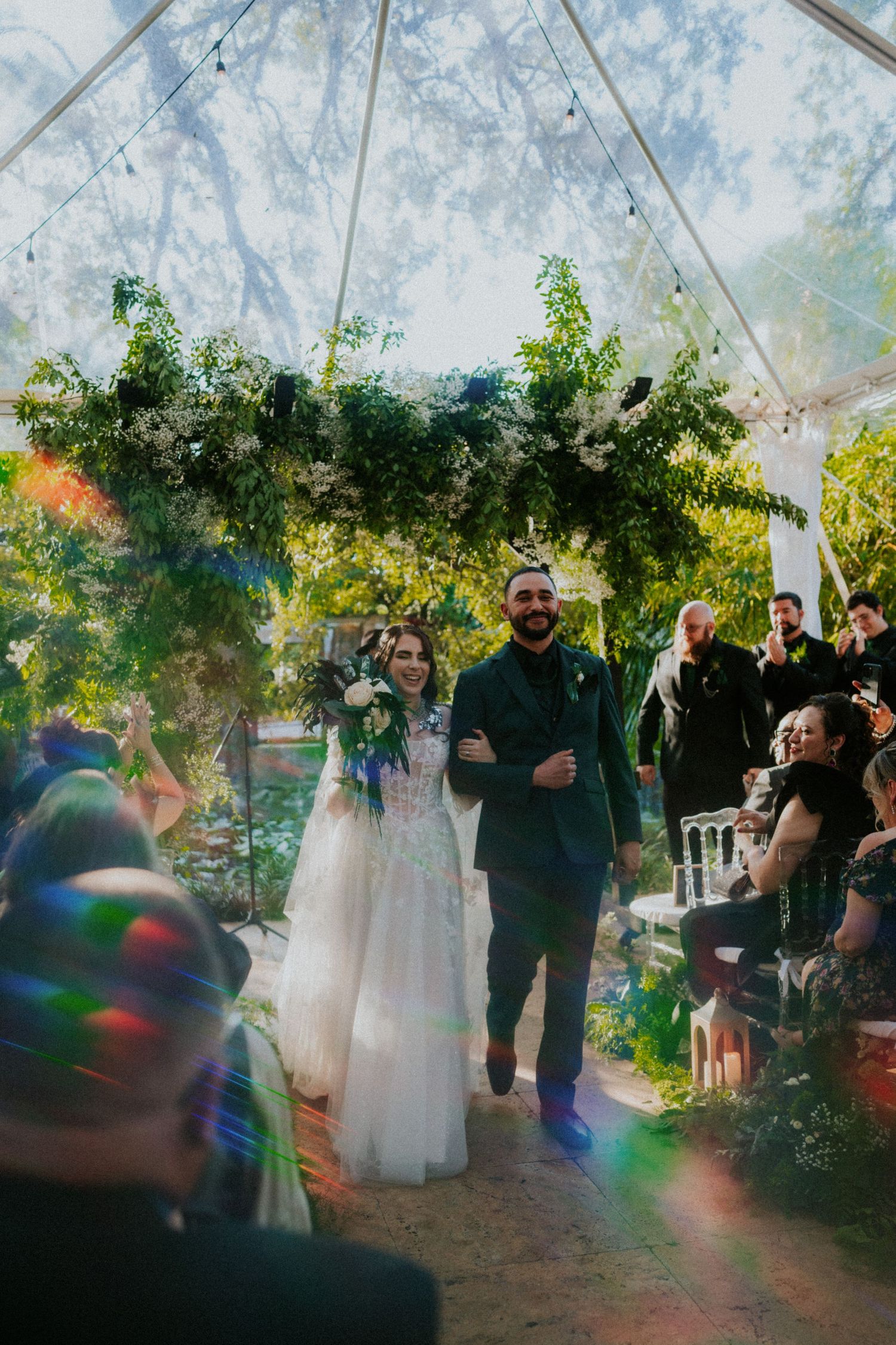 Joyful newlyweds walk down the aisle under a lush floral arch in a sunlit garden wedding ceremony.