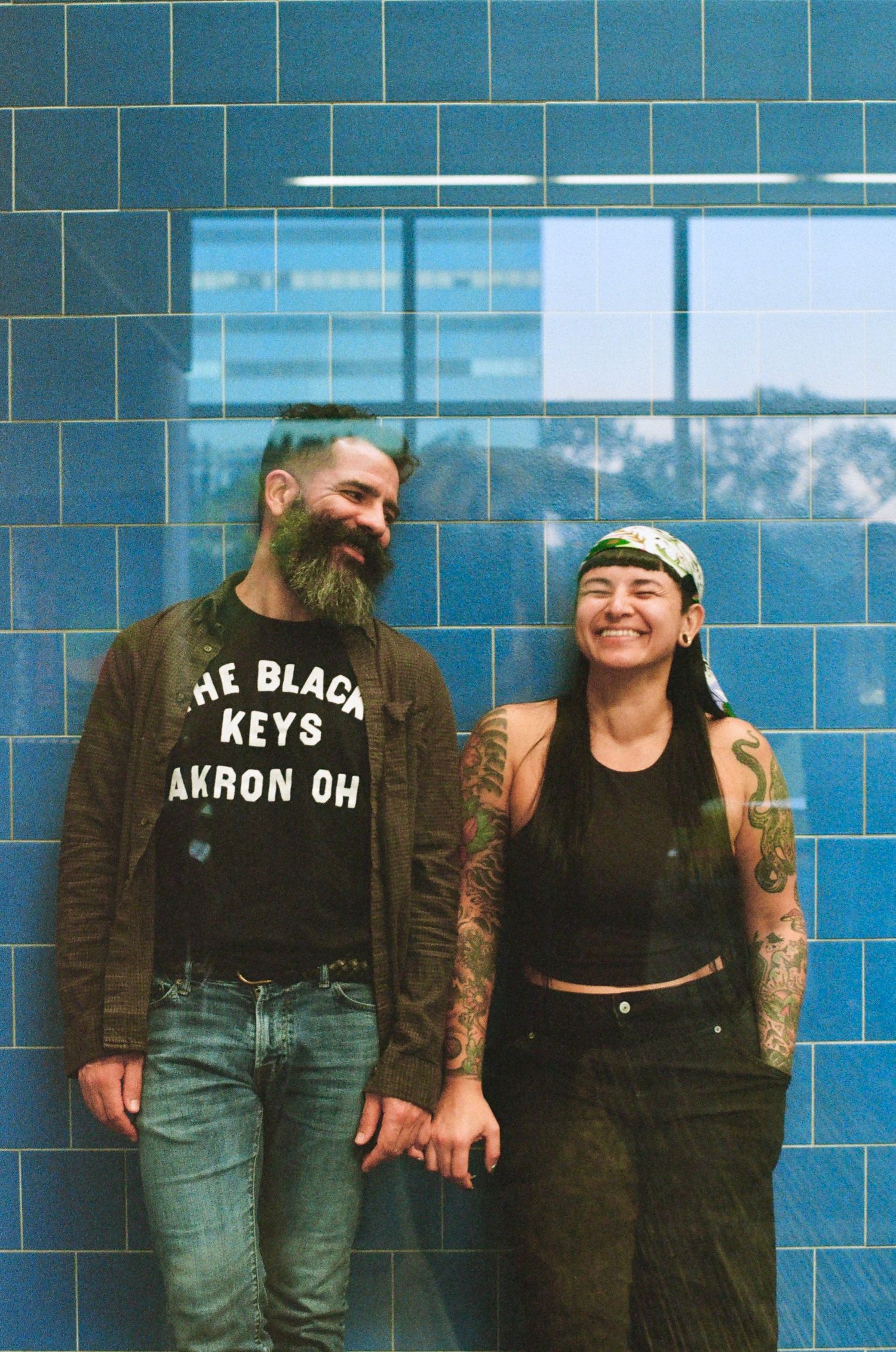 A bearded man in a Black Keys t-shirt and a tattooed woman in a bandana laugh together against a blue tiled wall.