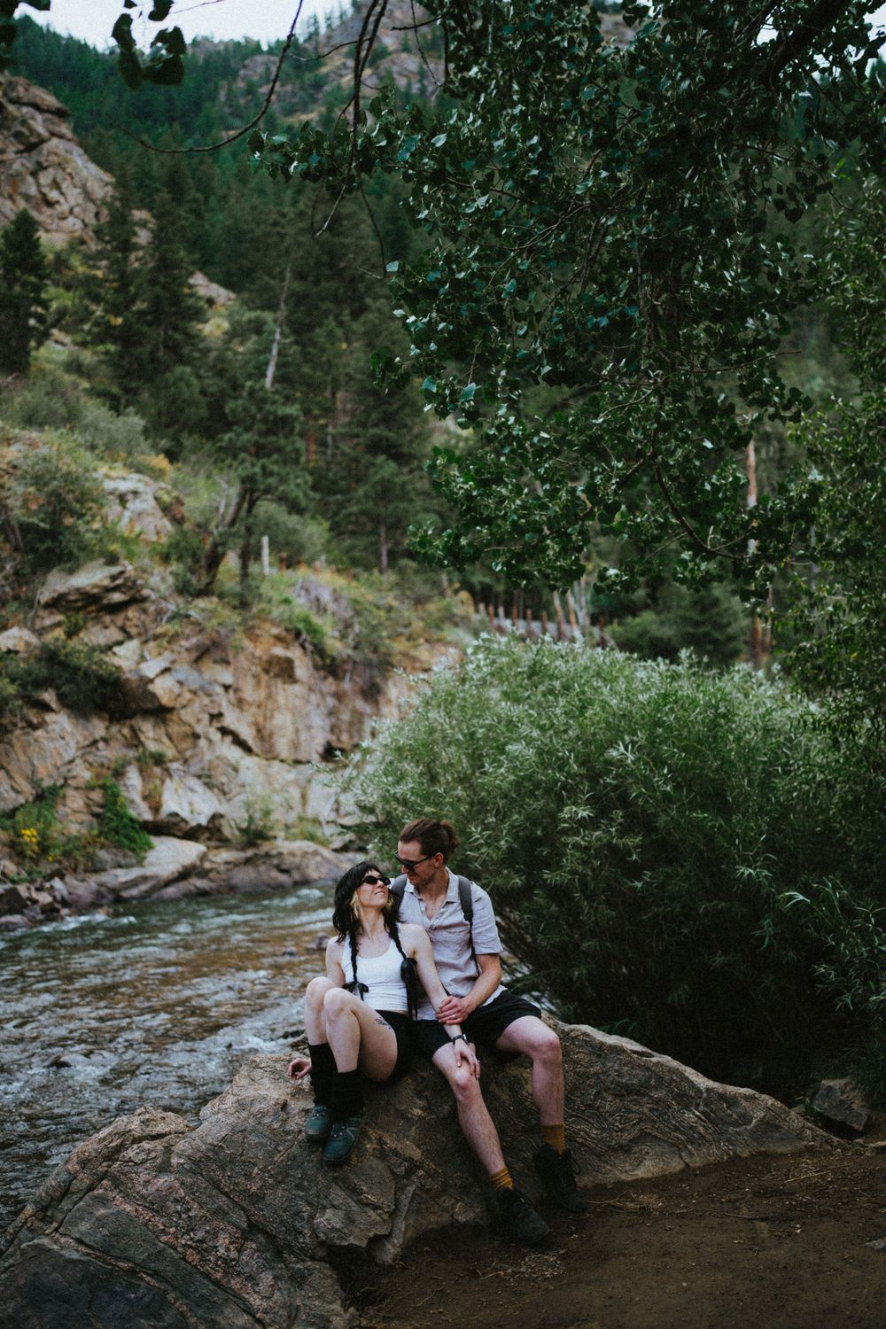 Two hikers resting on rocks by a creek surrounded by green foliage in the wilderness.