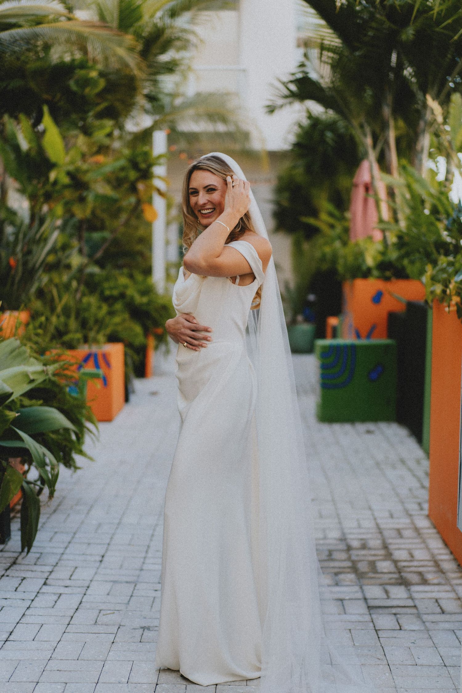 Elegant bride in white off-shoulder wedding dress and veil posing in tropical garden pathway.