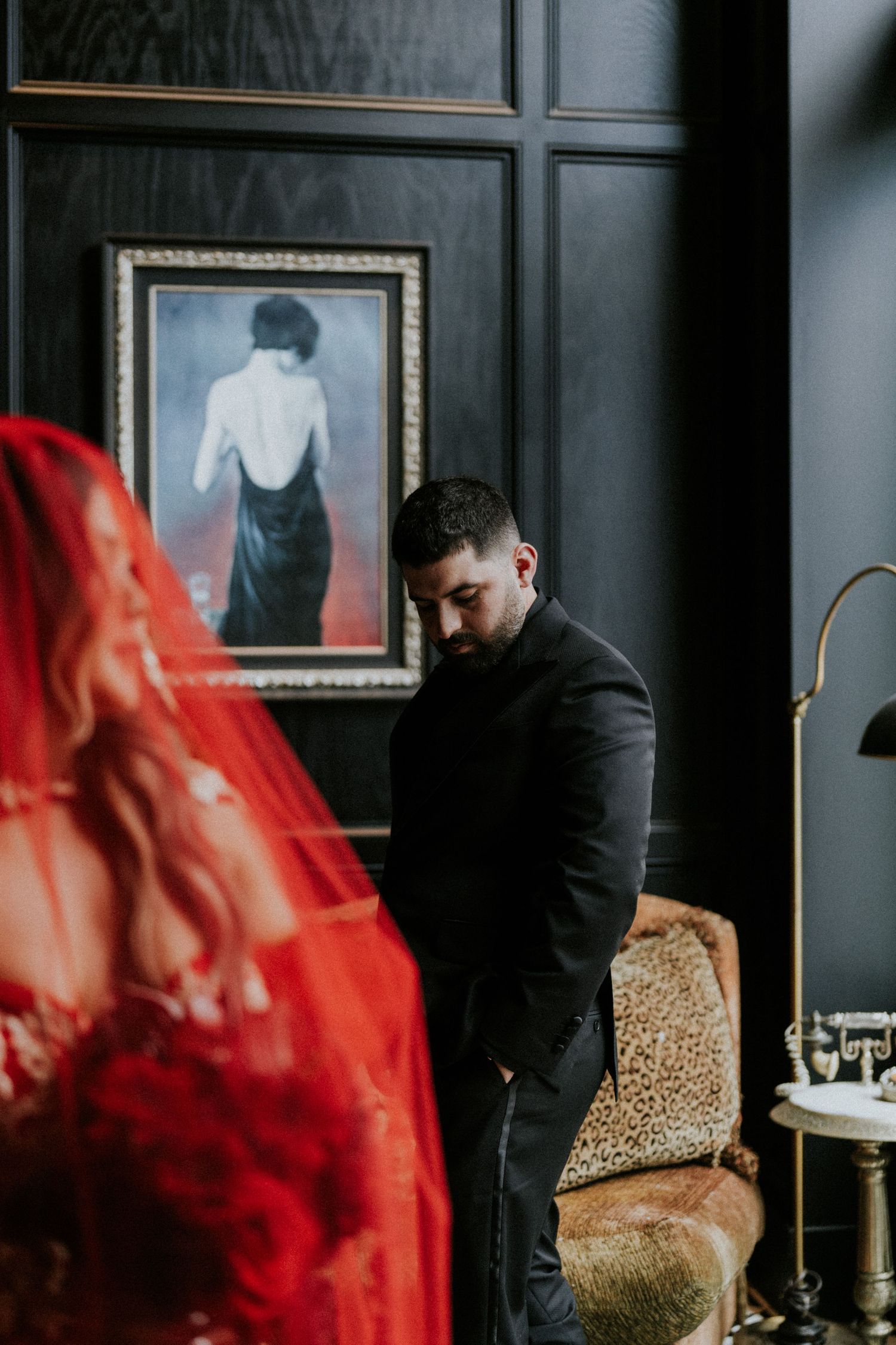 Man in black suit stands pensively near a painting, with a red veil in the foreground in a dark elegant room.