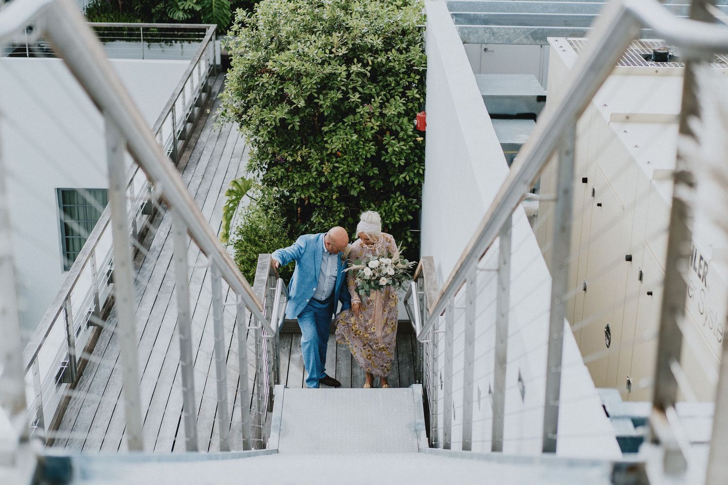 Couple walking up outdoor stairs with metal railings, surrounded by lush greenery, dressed in wedding attire.