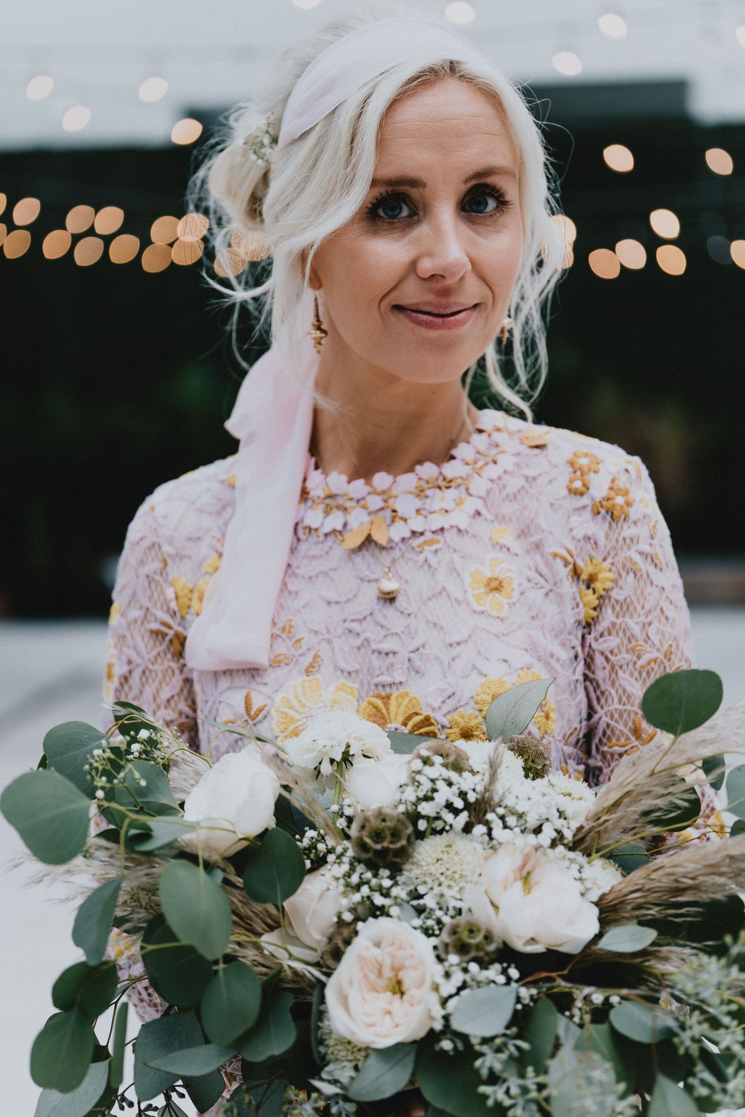 Blonde bride in pink floral lace dress holds white and green bouquet with fairy lights bokeh background.