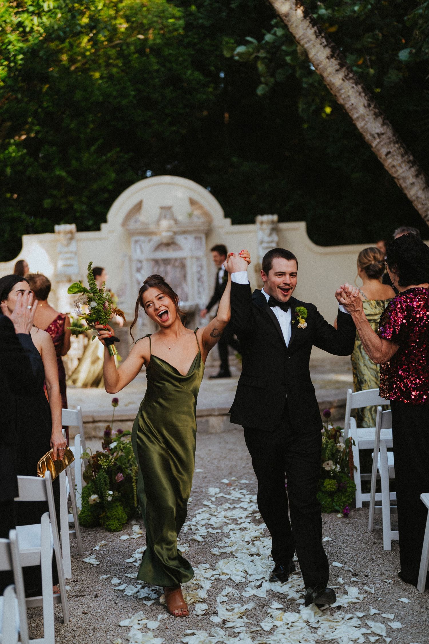 Joyful newlyweds walk back down the aisle at an outdoor garden wedding ceremony with white petals on the ground.