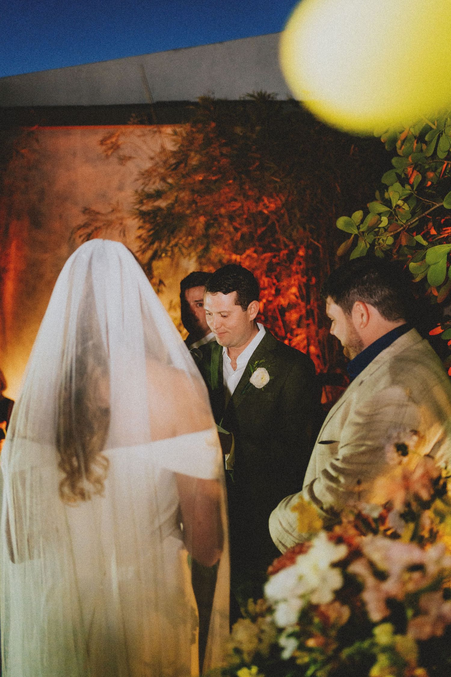 Groom gazes at veiled bride during an intimate outdoor wedding ceremony at dusk.