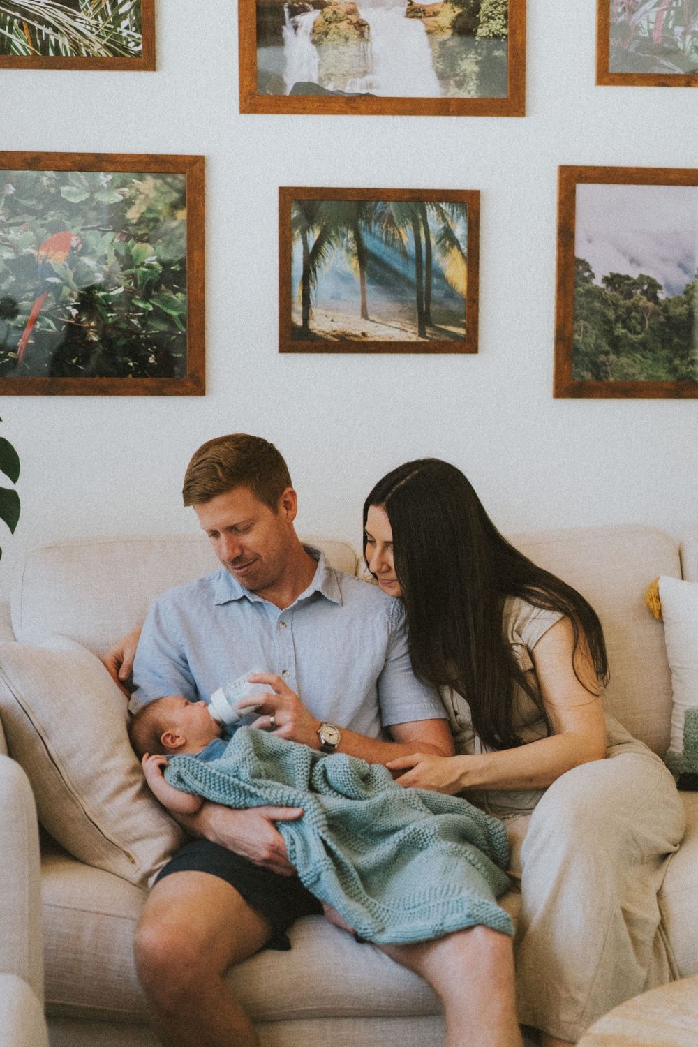 A couple sits on a beige couch feeding an infant wrapped in a blue blanket, with framed artwork on the wall behind them.