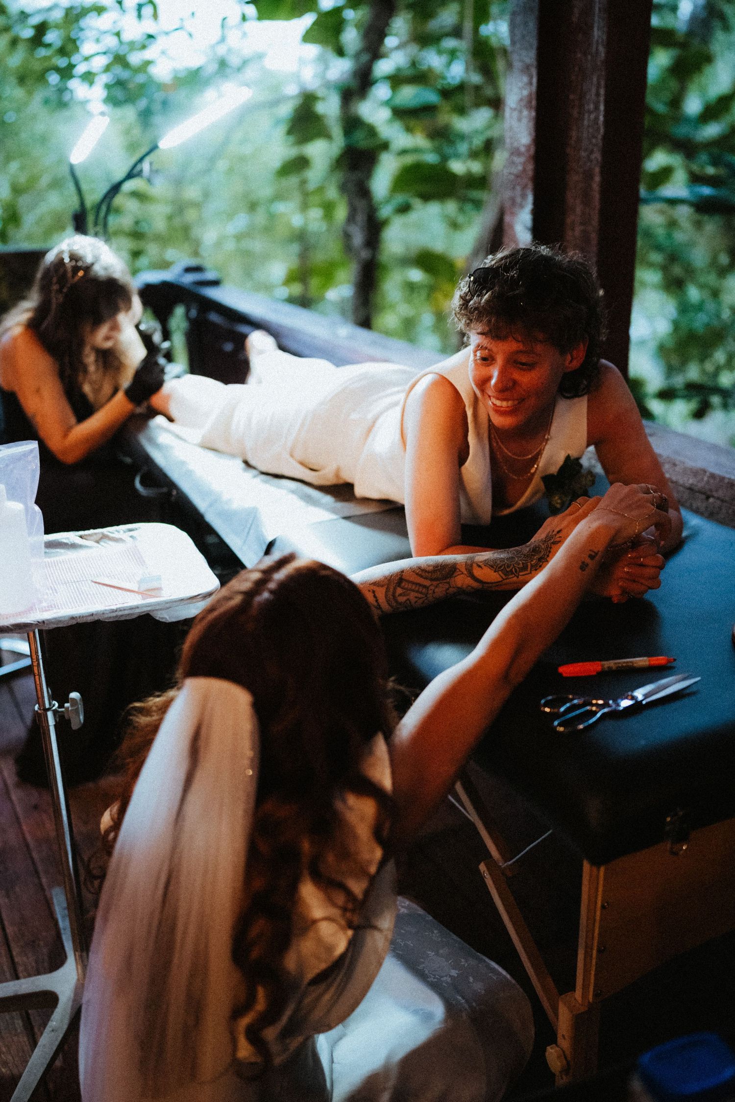 Bride in veil holds groom's tattooed hand as he lies on massage table getting a tattoo on their wedding day.