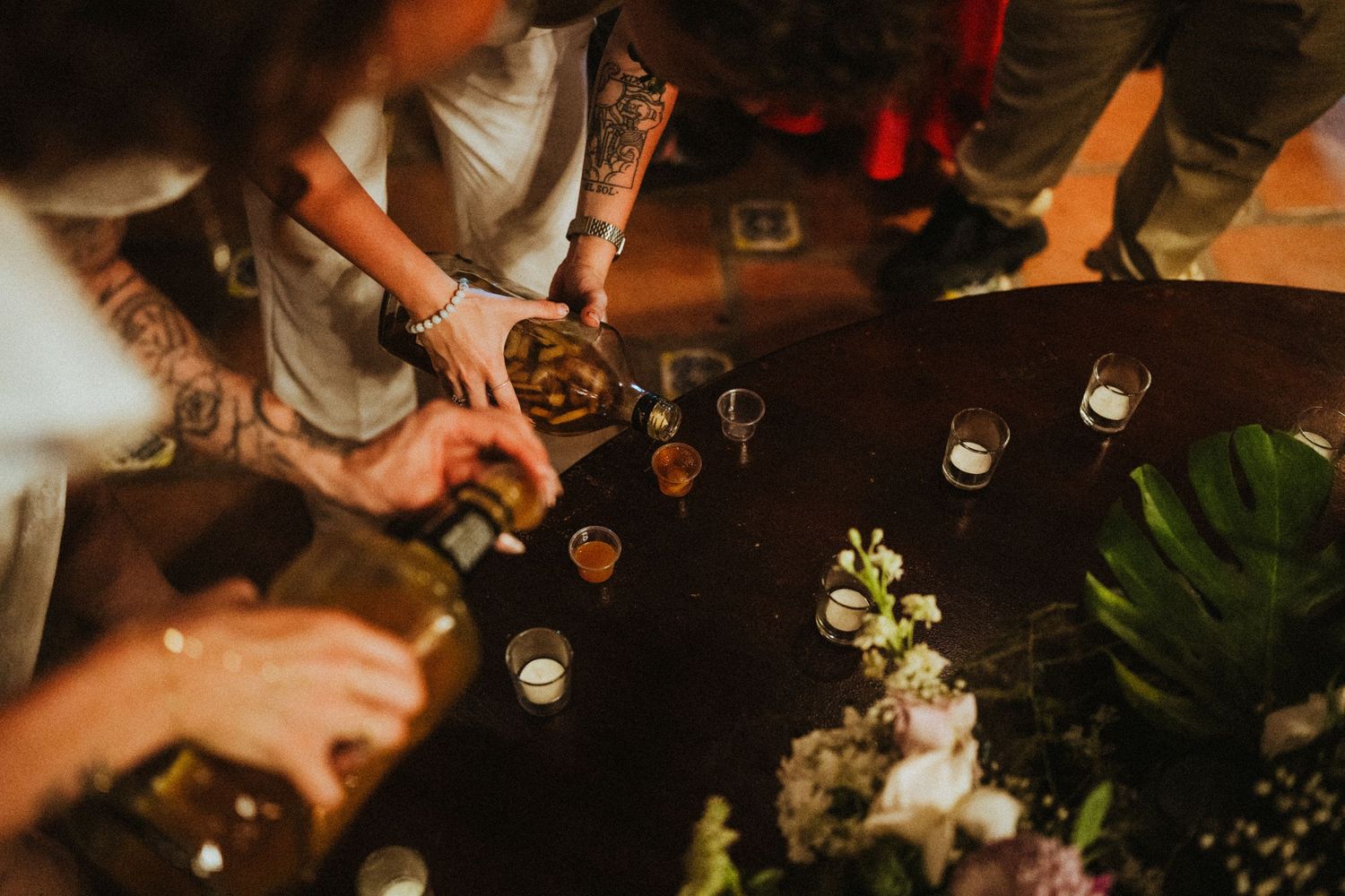 Aerial view of tattooed guests pouring drinks at a round table with floral centerpiece and candles at a party.