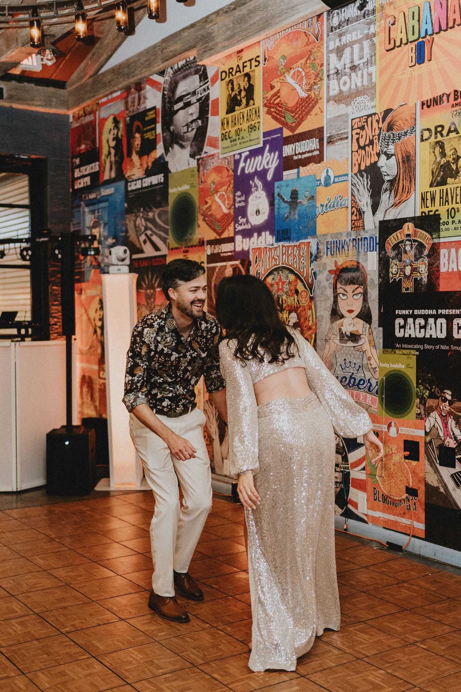 Joyful couple dances together in front of a colorful concert poster wall, bride wearing a sparkling sequin two-piece gown.