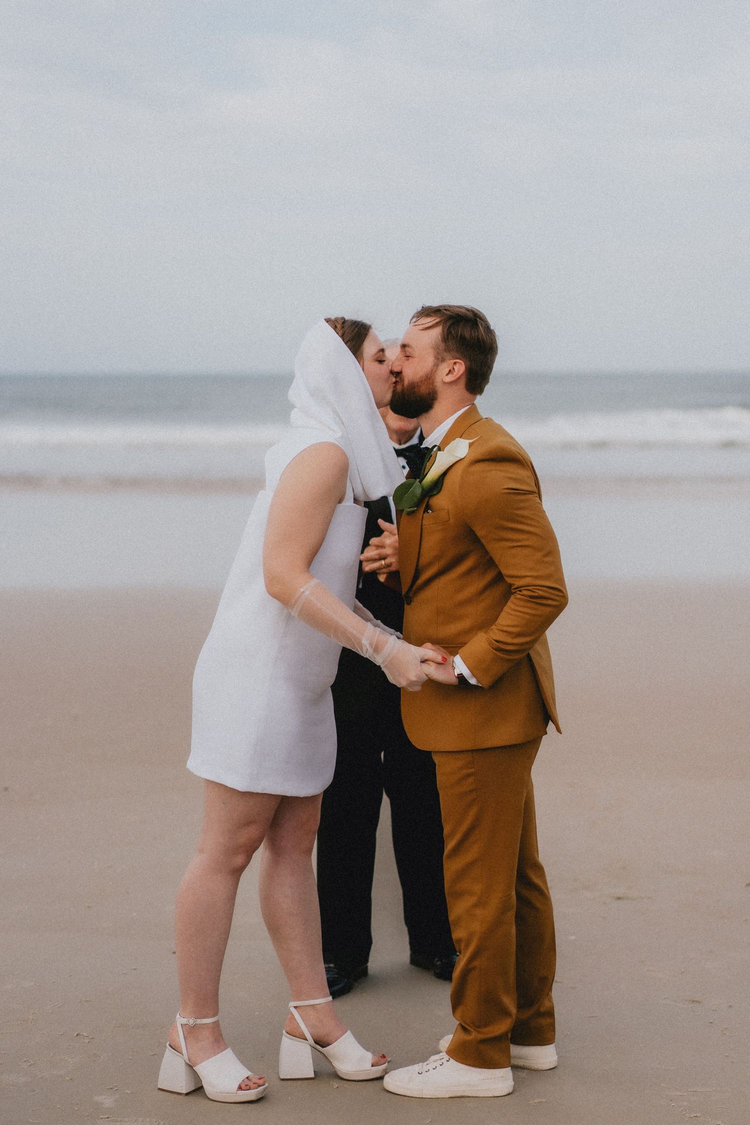Newlyweds share first kiss on sandy beach, bride in white mini dress, groom in mustard suit.