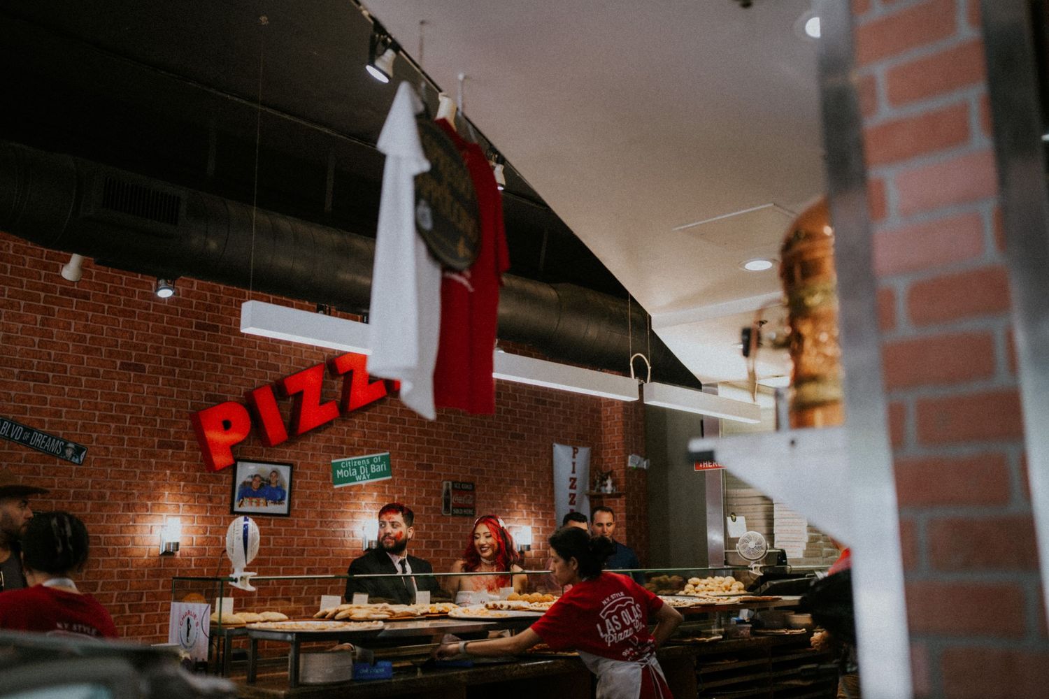 Interior of a rustic pizza restaurant with brick walls, red PIZZA sign, and diners seated at the counter.