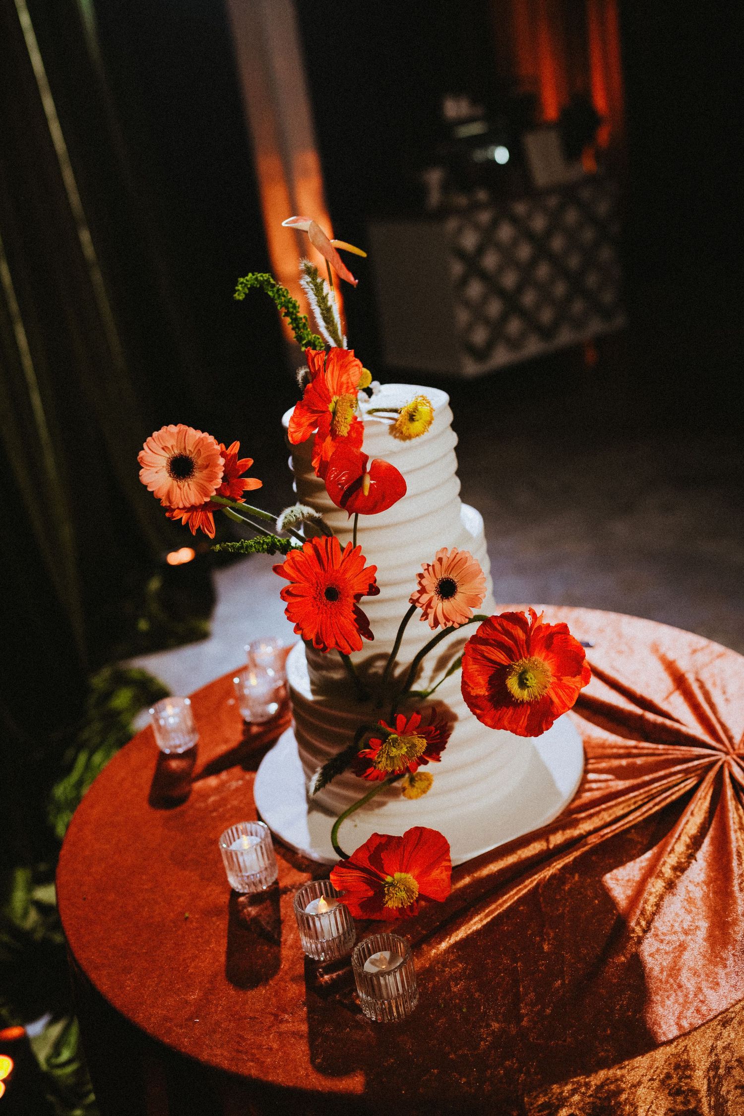 White tiered wedding cake adorned with vibrant orange gerbera daisies and poppies on a copper sequin table.