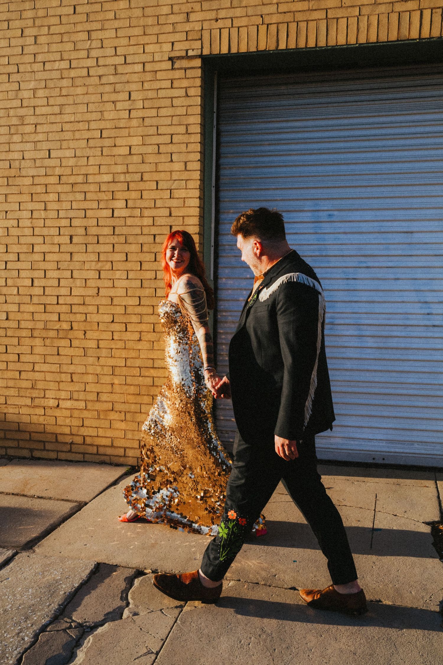 Bride in gold sequin dress and groom in black suit walk hand-in-hand on a sunlit urban sidewalk.