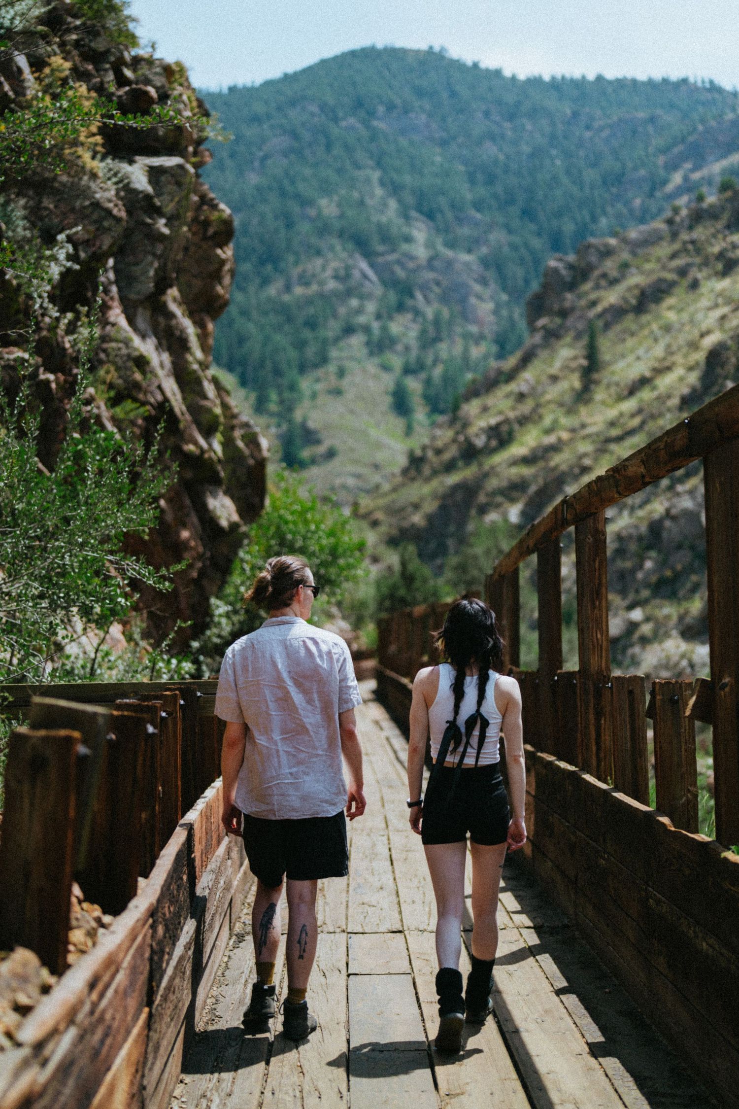 Couple walking on wooden bridge trail through mountain canyon with lush green trees and rocky cliffs.