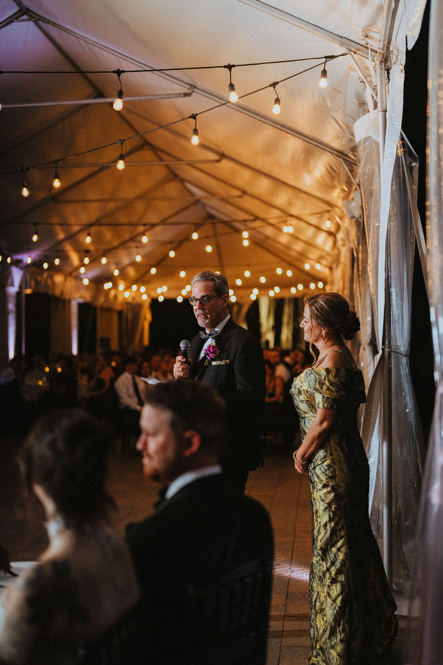 Man giving speech at elegant tented wedding reception with string lights while woman in floral gown stands nearby.