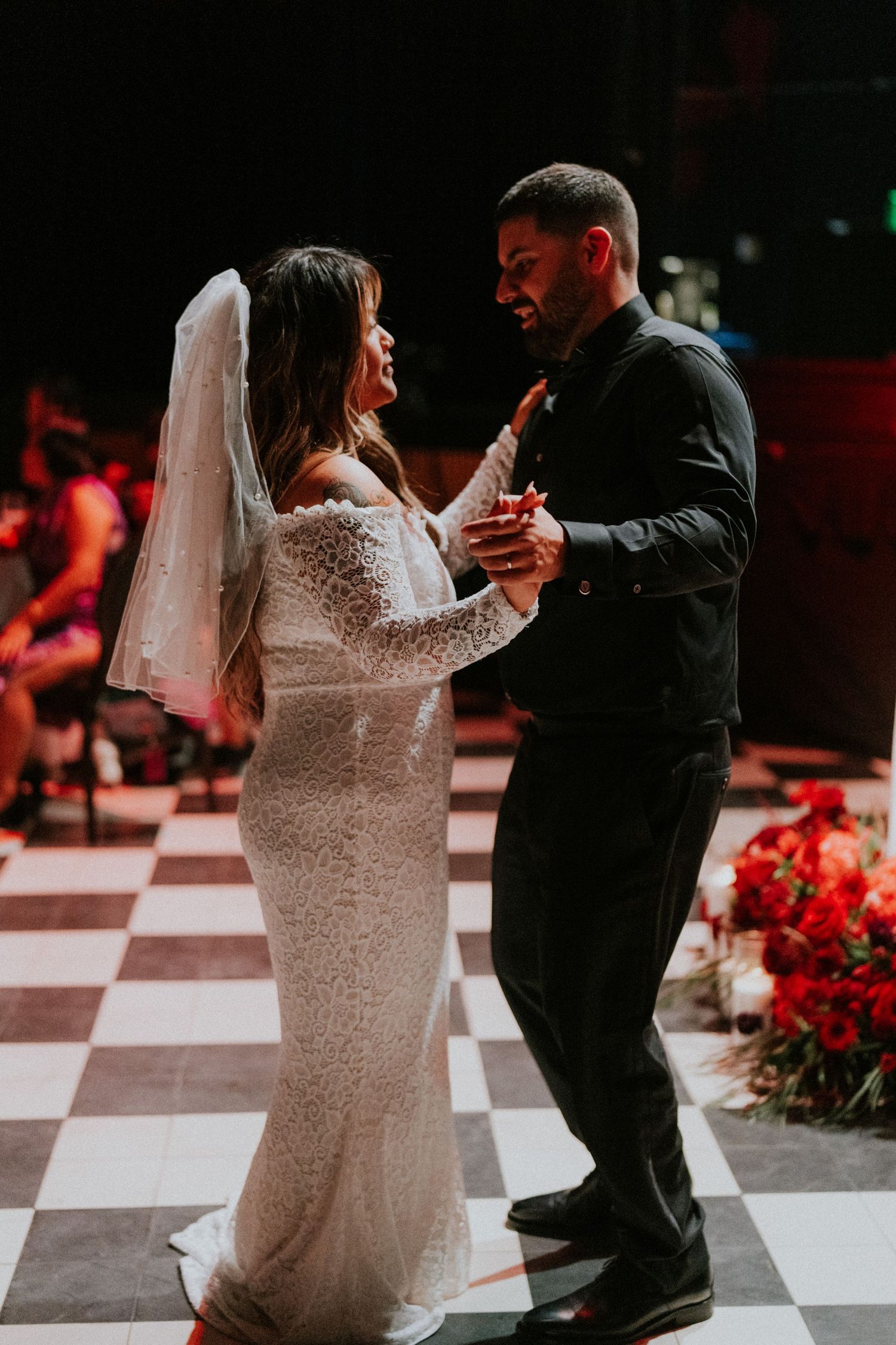 Bride and groom share their first dance on a checkered floor, surrounded by red floral arrangements at their wedding reception.
