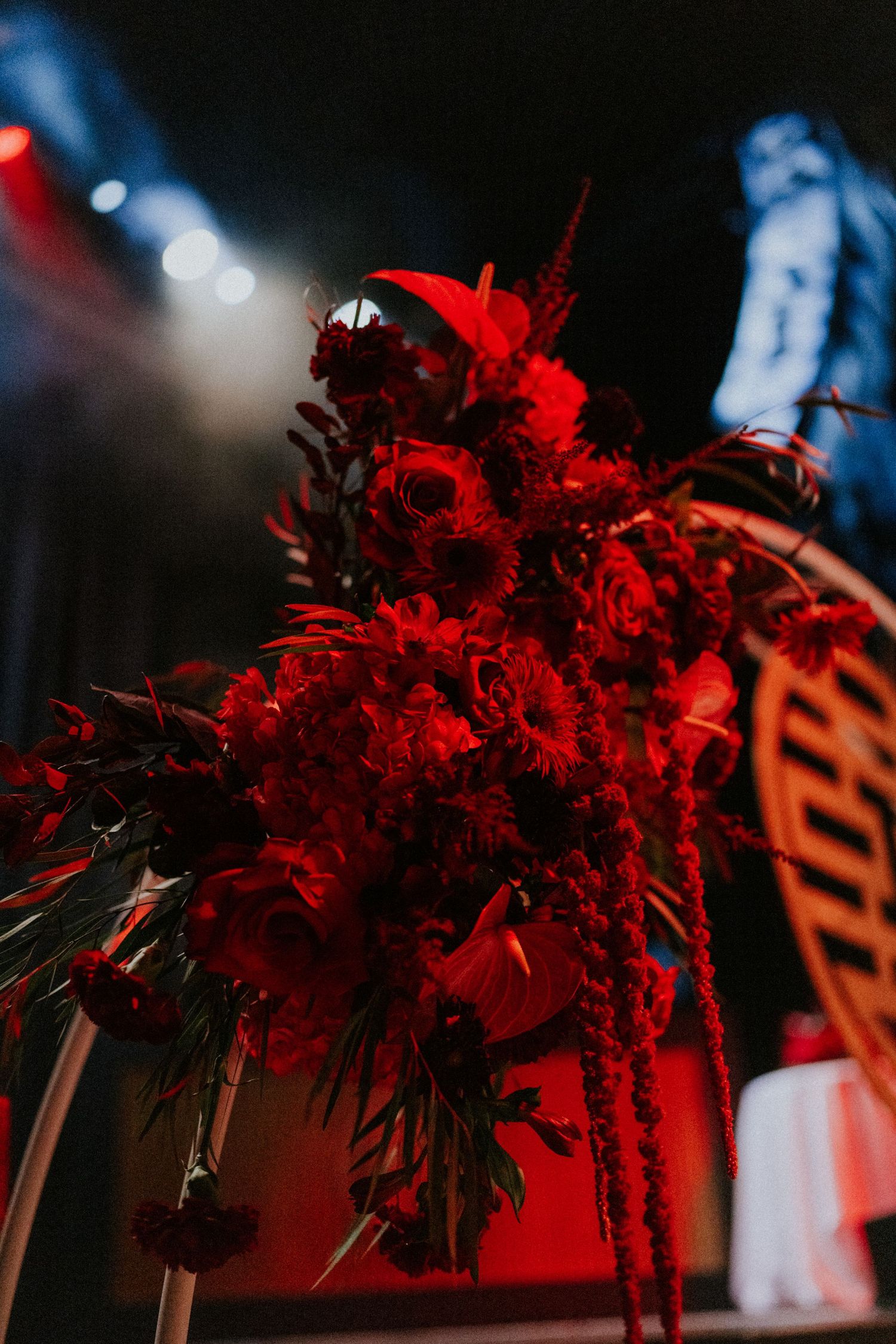 Dramatic red floral arrangement with roses and feathers displayed on a stand under moody red stage lighting.