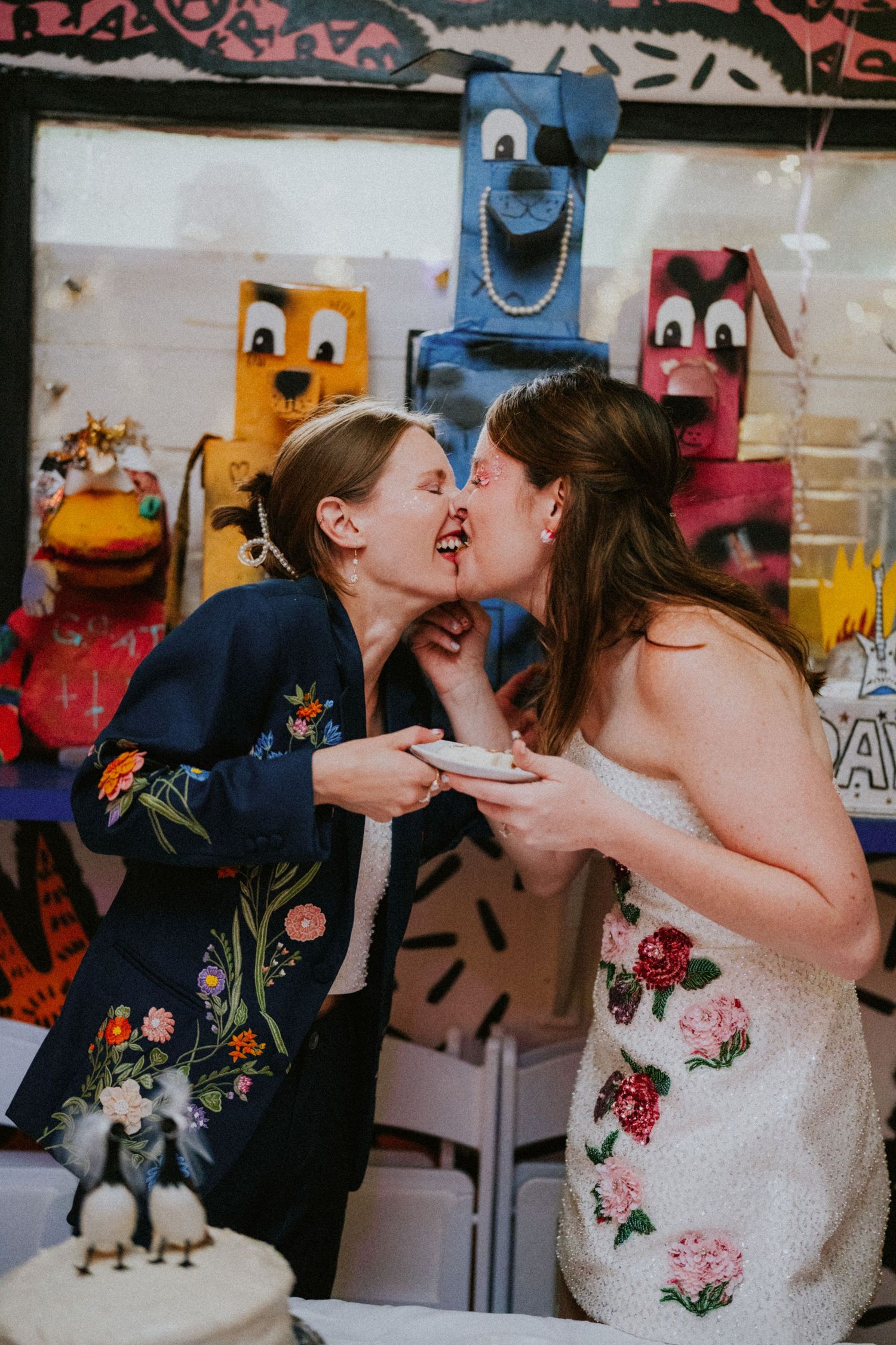 Two brides share a playful cake-tasting kiss at their whimsical wedding reception with colorful puppet decor.