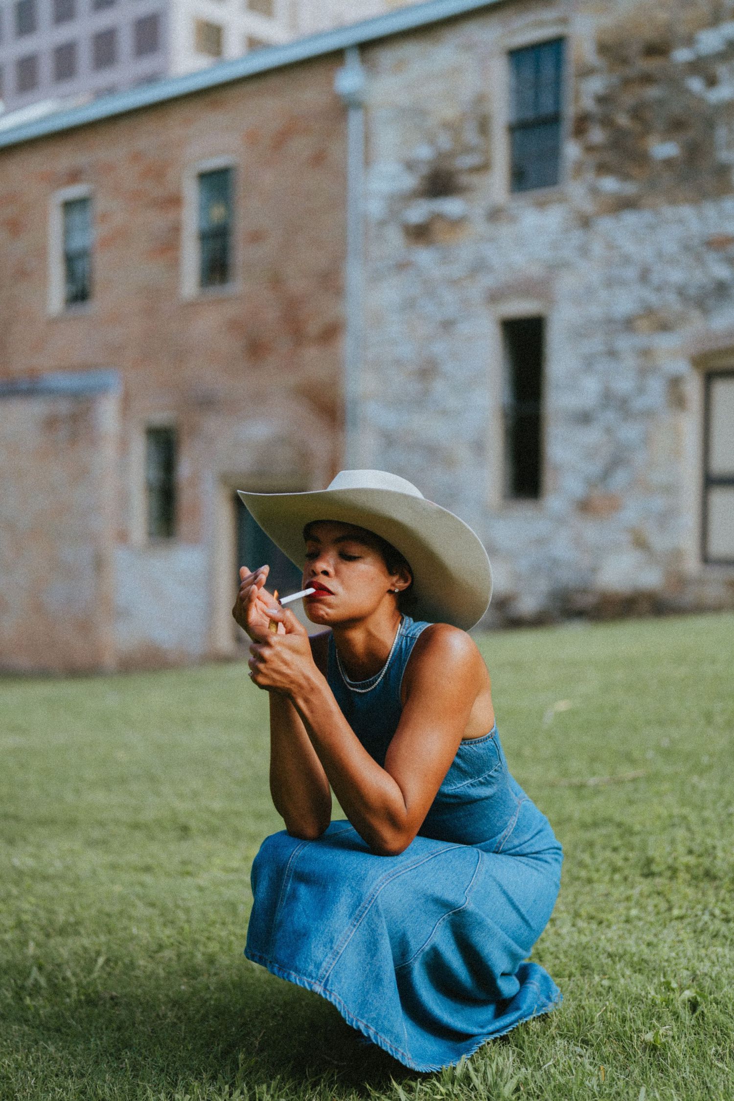 Woman in blue dress and wide-brim hat crouching on grass, lighting a cigarette near a rustic stone building.