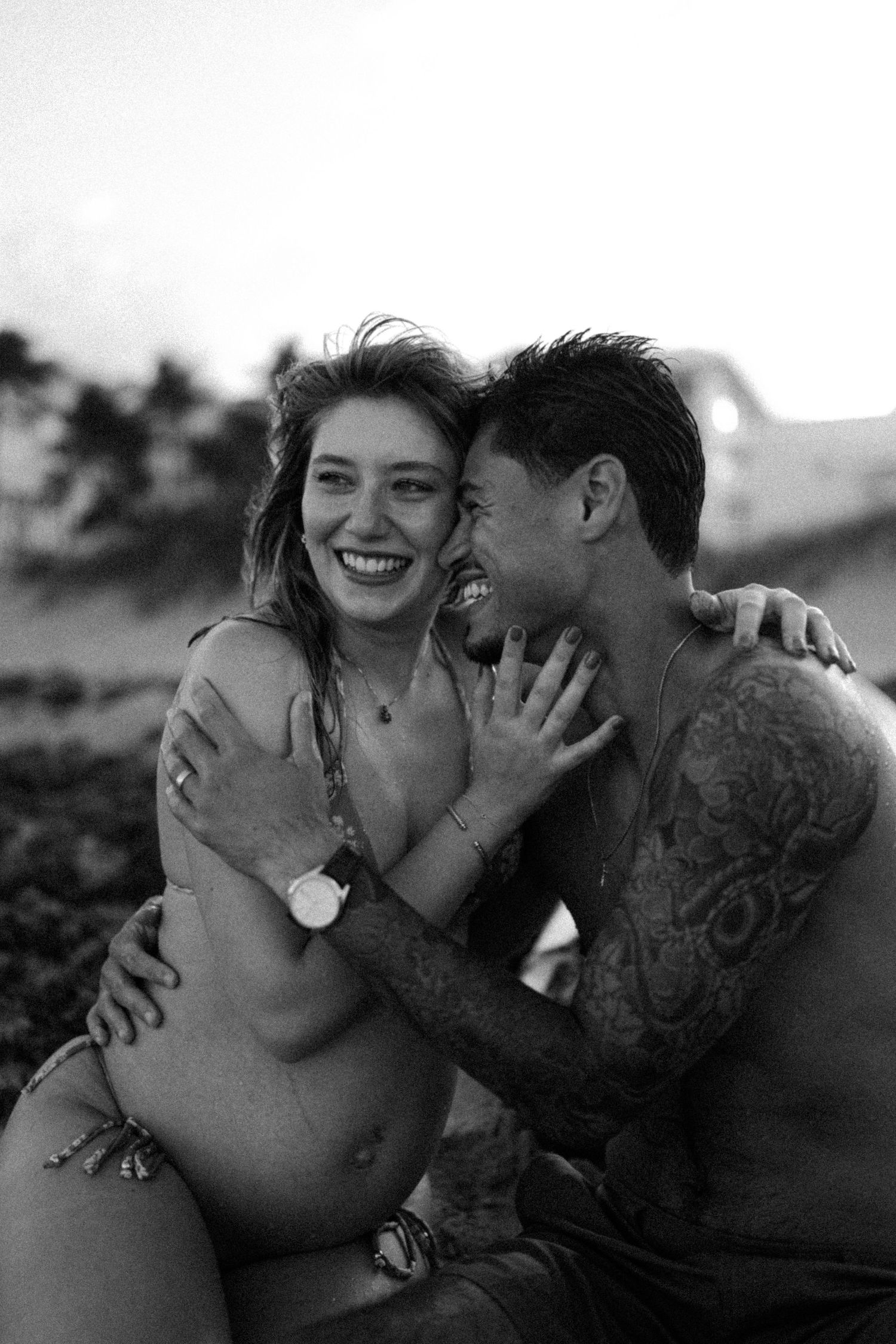 Black and white photo of a laughing couple embracing on a beach, man with tattoos hugging smiling woman.