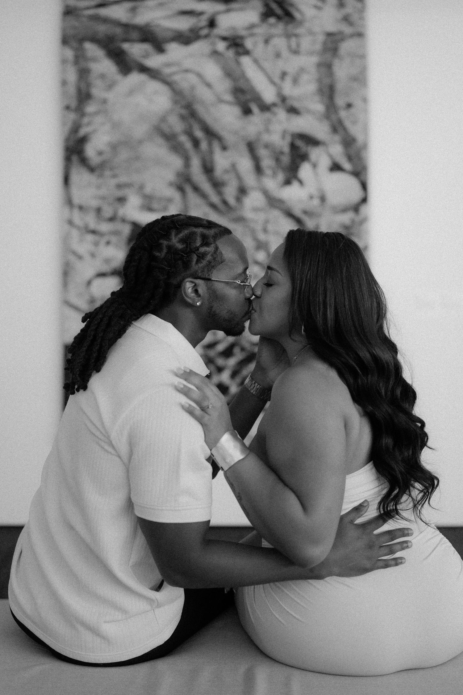 Black and white photo of a couple sharing a romantic kiss while sitting on the floor in front of an abstract painting.