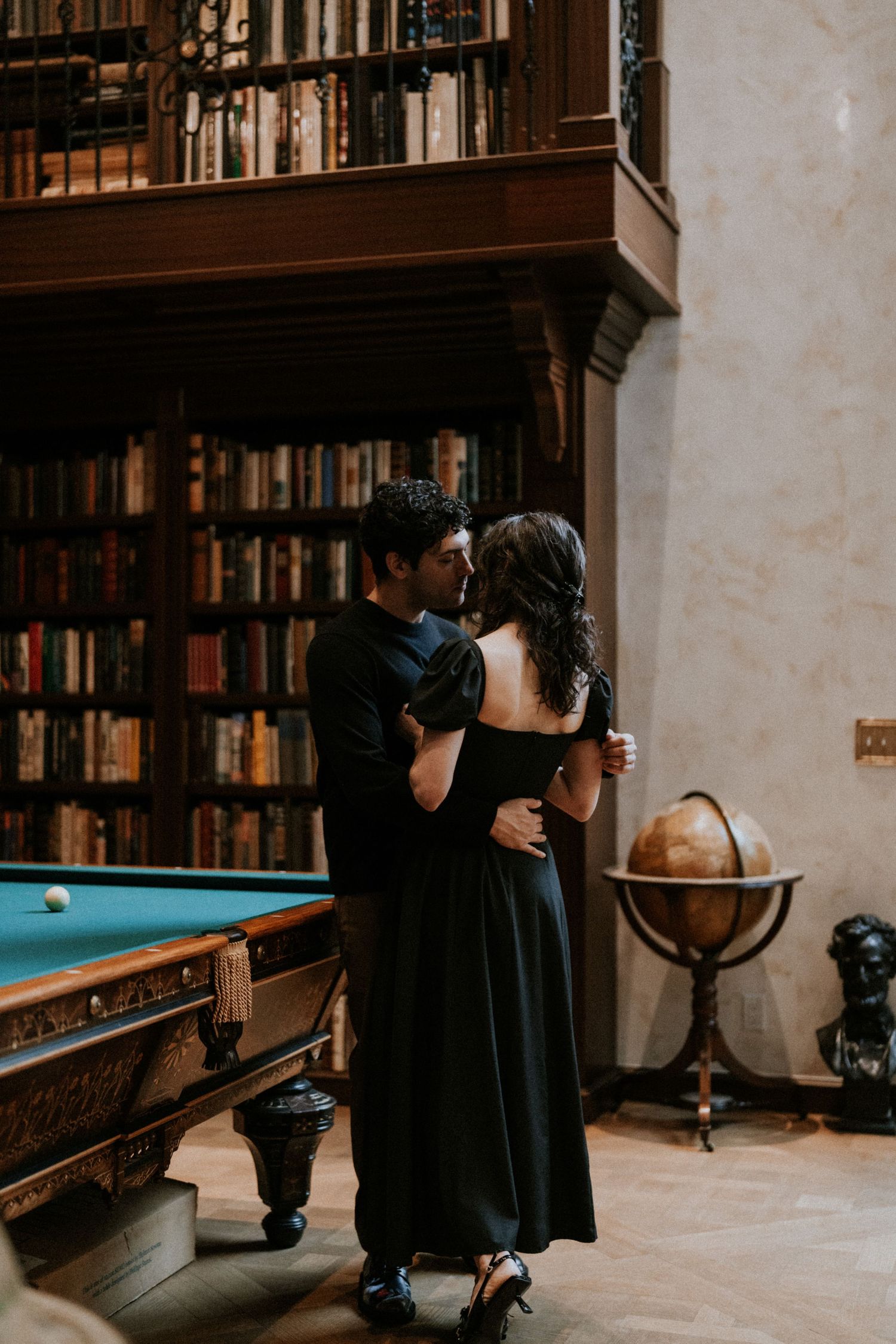 Couple in black attire embracing romantically in an elegant library with bookshelves, billiard table, and vintage globe.
