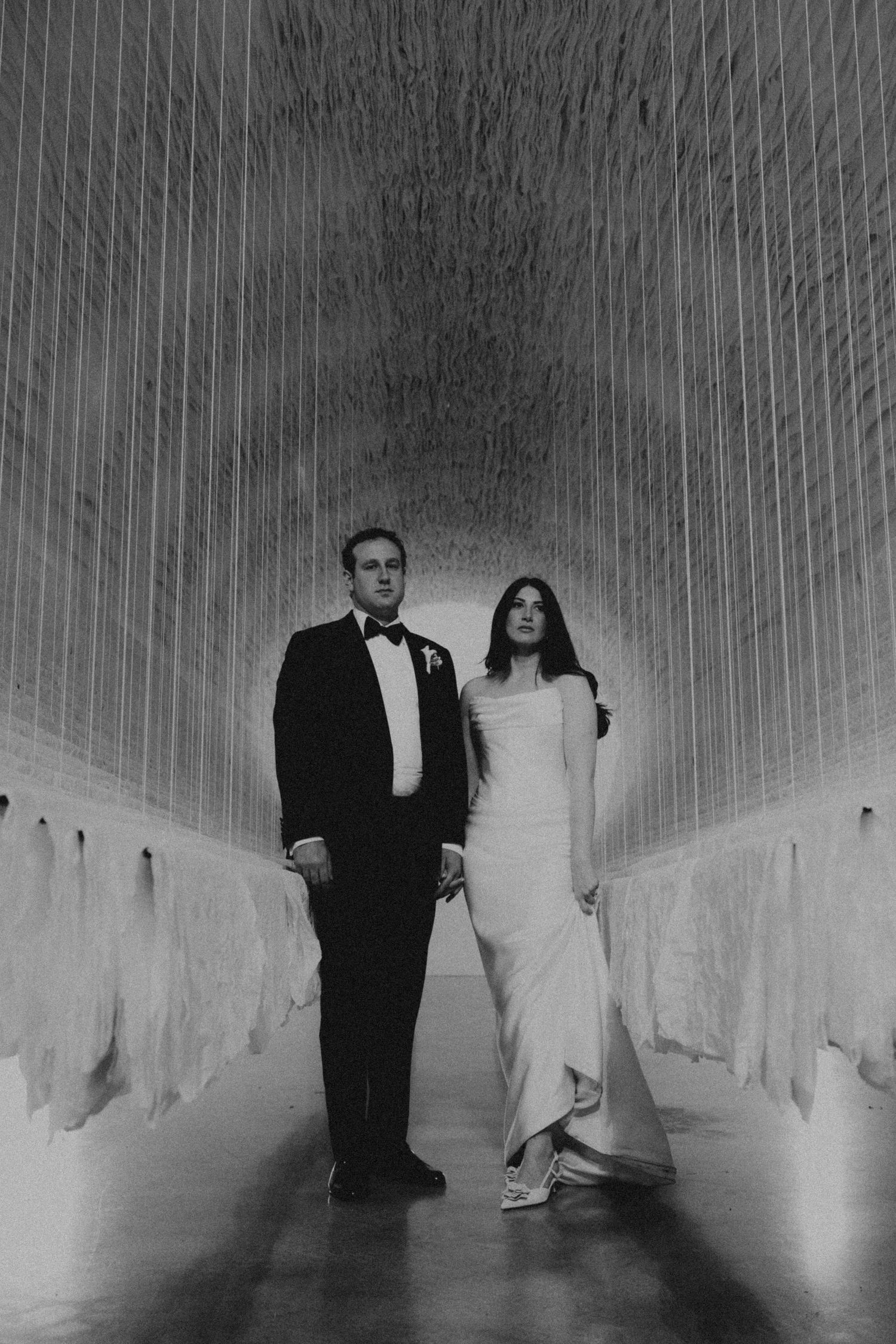 Black and white photo of a bride and groom walking through an elegant tunnel adorned with hanging fabric at their wedding.