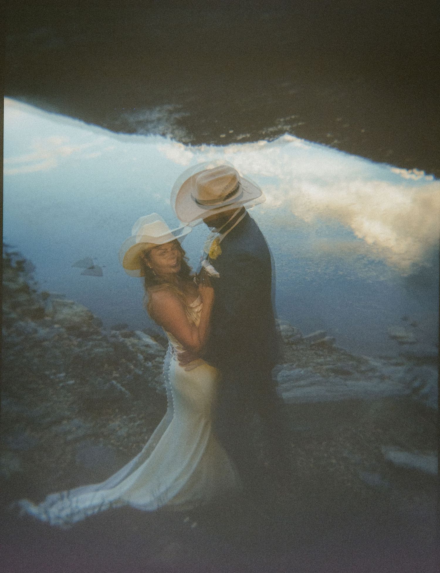 A couple in cowboy hats embraces in a dreamy double-exposure wedding photo blending sky and landscape.