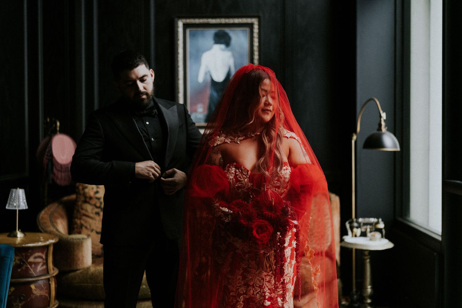 Bride in red wedding gown and veil with groom in black suit in a dark moody room.