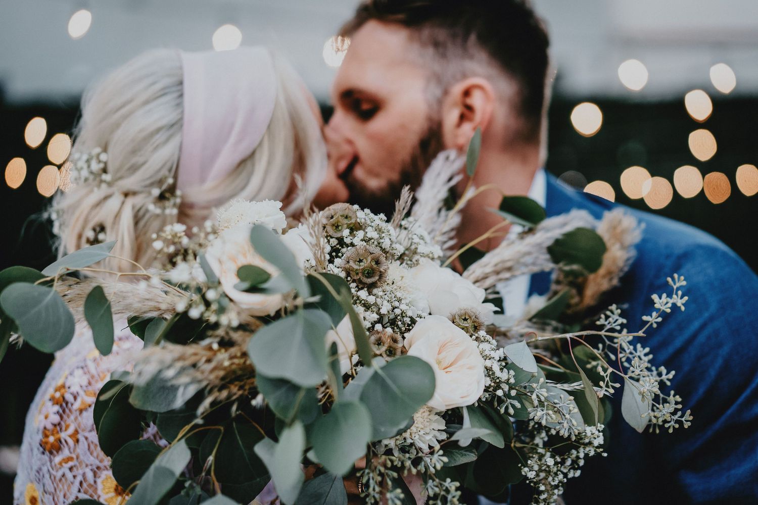 Couple sharing a romantic kiss behind a lush wedding bouquet of eucalyptus and white flowers with bokeh lights.