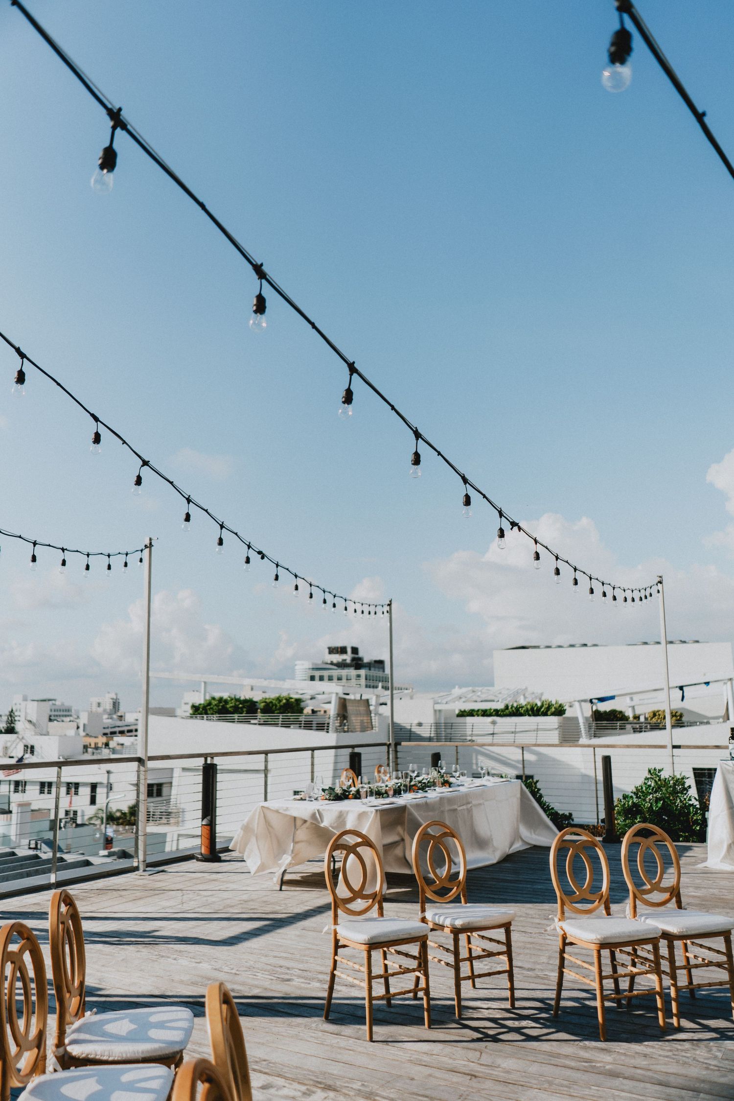 Rooftop wedding reception with string lights, elegant white tablecloth, gold chairs, and a clear blue sky backdrop.