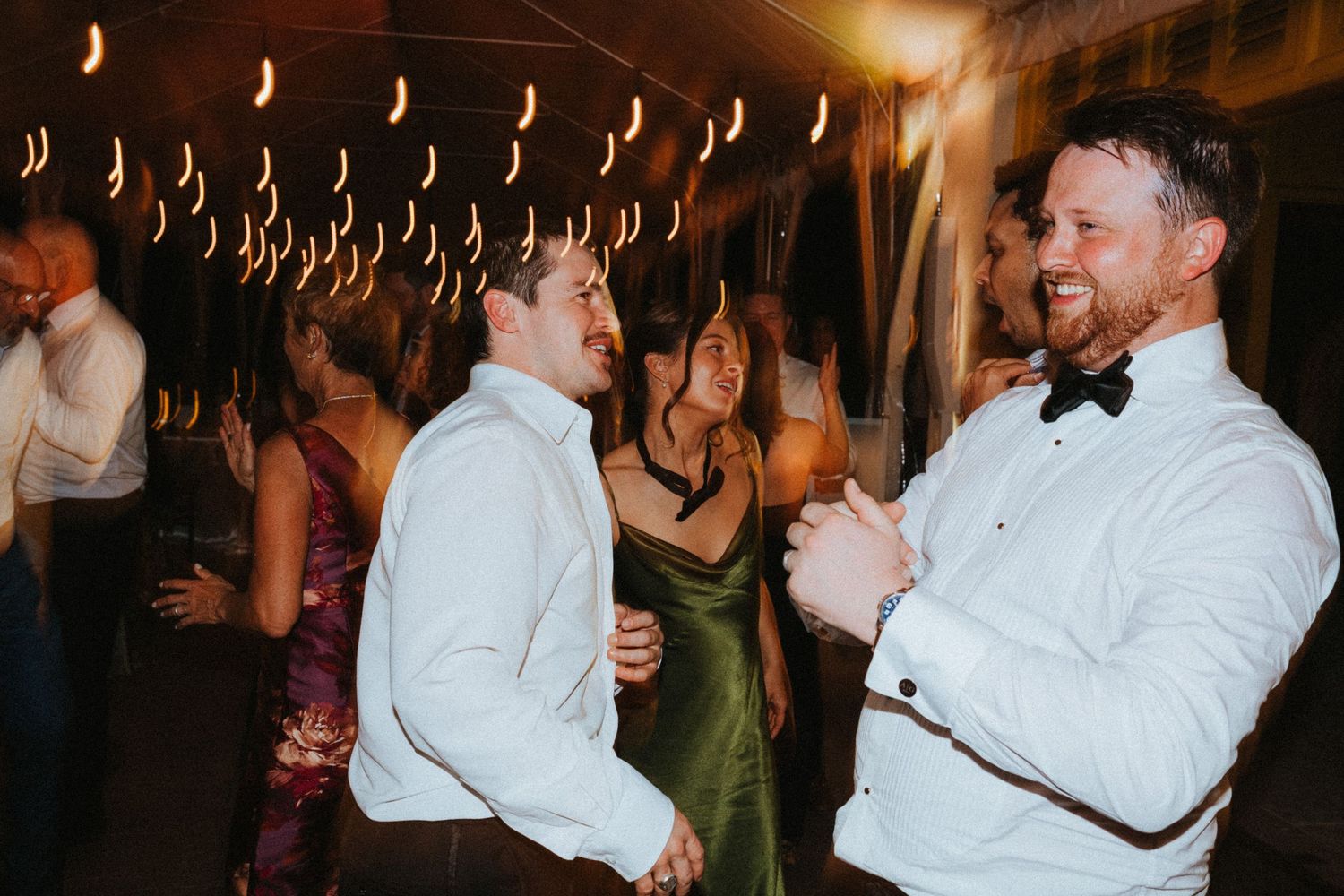 Two men in formal attire laugh and dance together at an elegant wedding reception on a dimly lit dance floor.