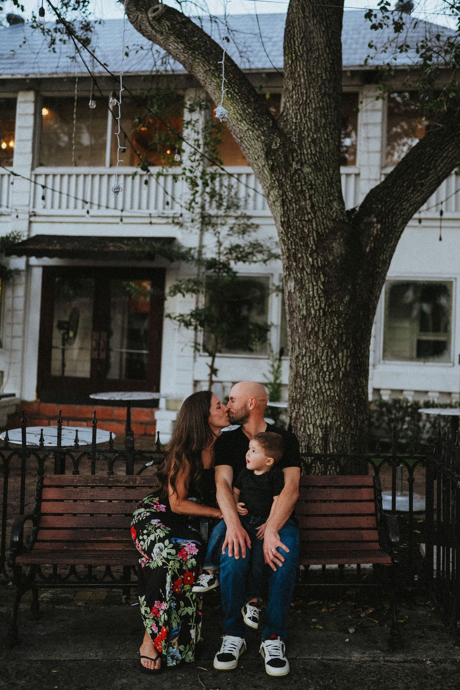 A couple shares a romantic moment on a wooden bench outside a charming white colonial-style house at dusk.