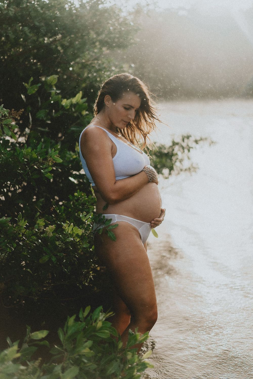 An expectant mother in white swimwear poses at sunset by the water during a maternity photoshoot.