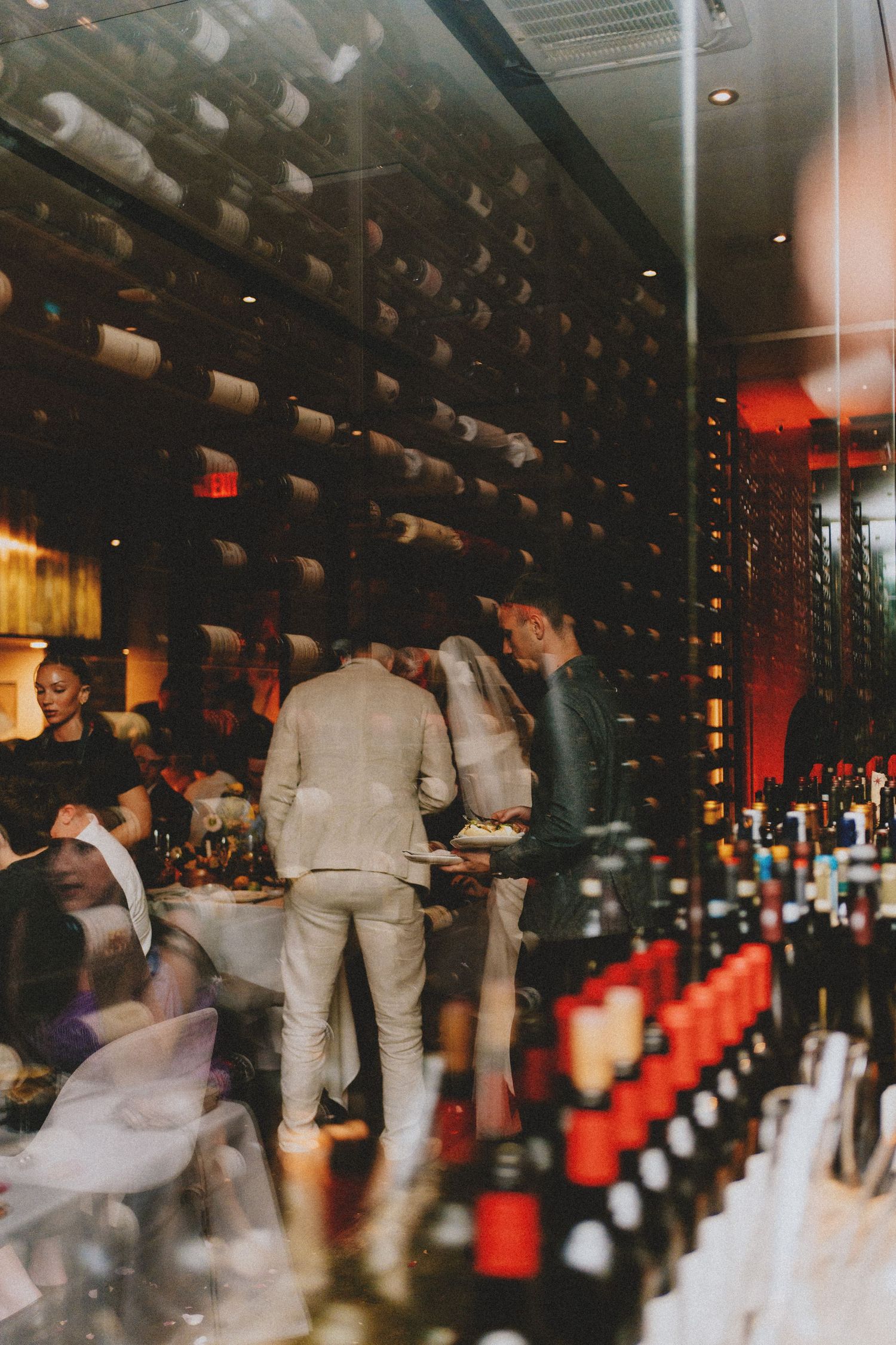 Wedding guests mingle near wine bottle display wall viewed through reflective restaurant window.