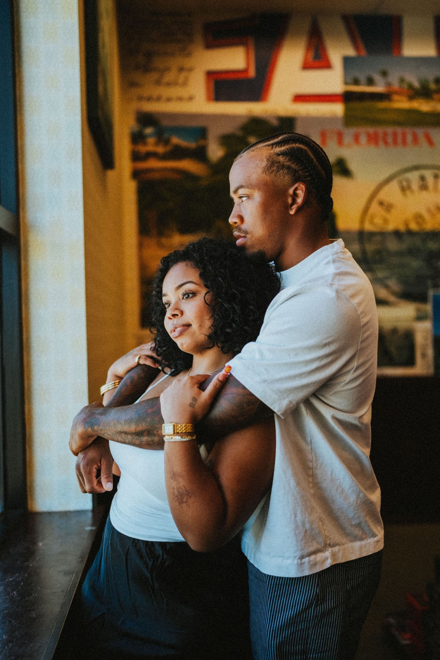 A couple embraces tenderly by a window, the man wrapping his arms around the woman from behind in warm light.