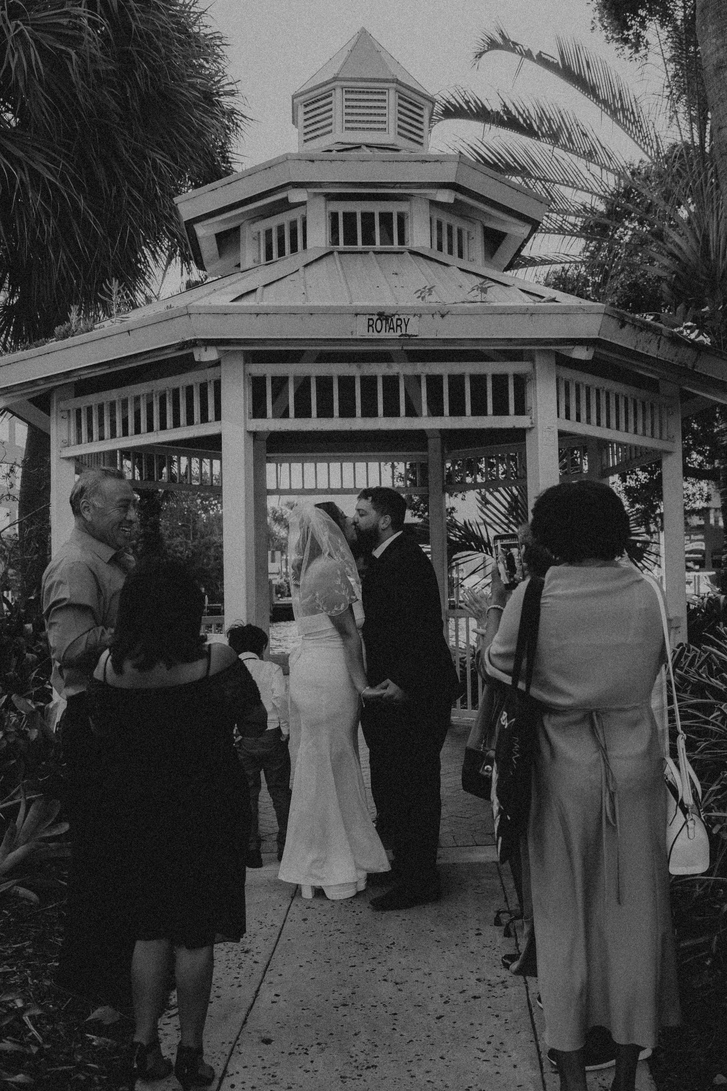 Black and white photo of a bride and groom kissing in a gazebo surrounded by wedding guests.