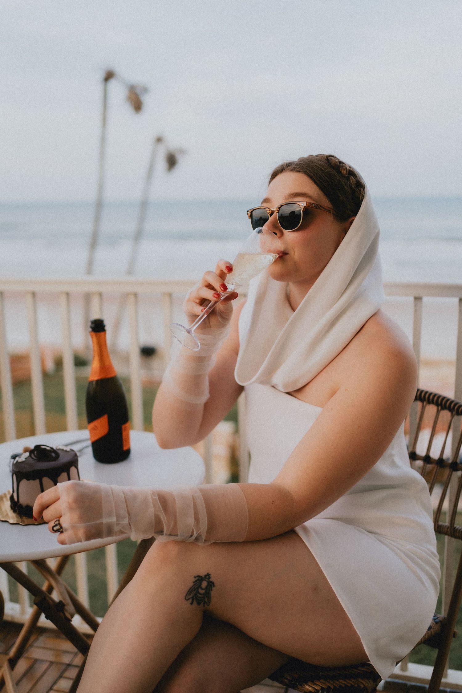 Bride in white dress and veil sips champagne on ocean-view balcony wearing sunglasses with cake nearby.