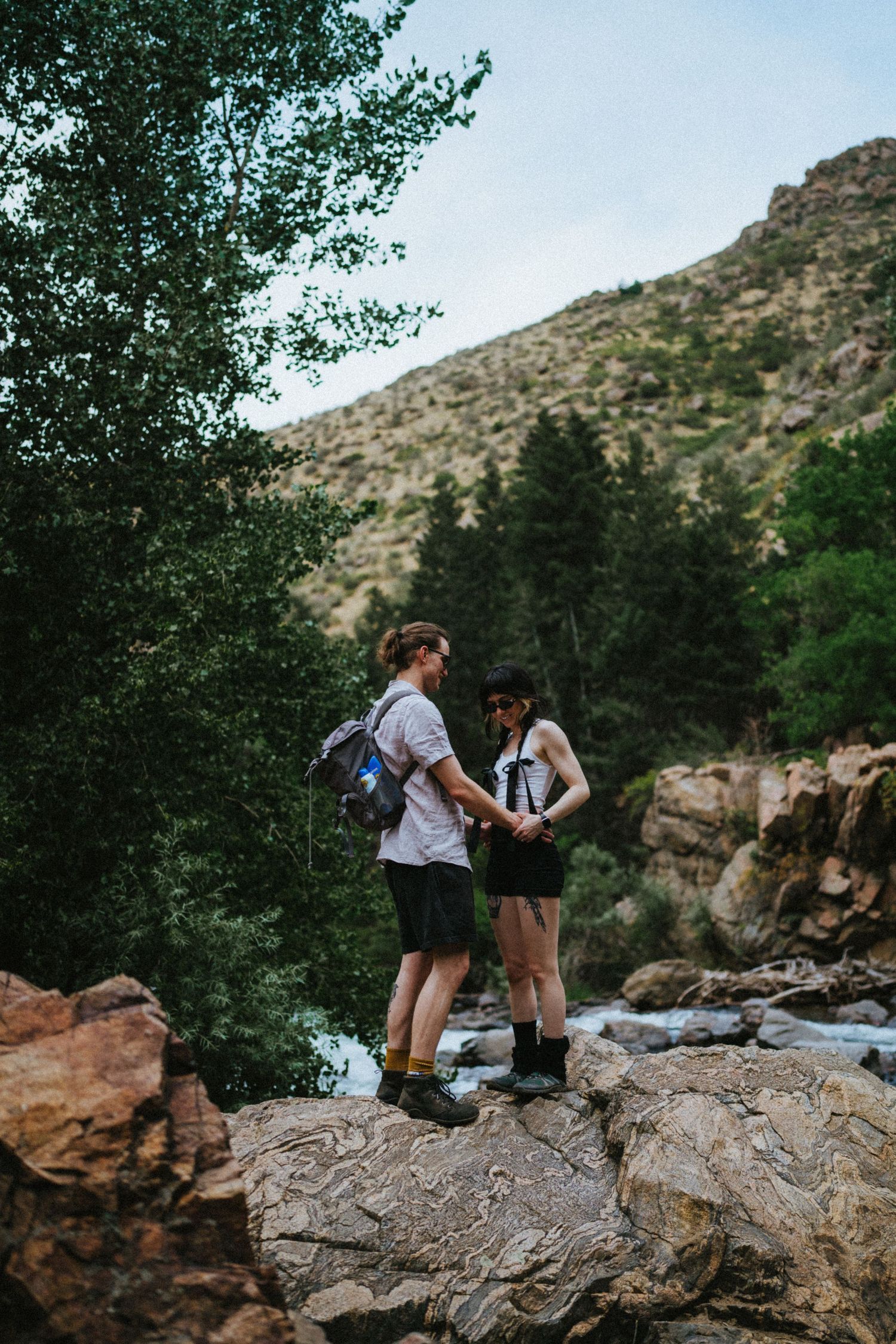 Two hikers with backpacks stand on rocks by a mountain stream surrounded by lush green trees and rocky hillside.