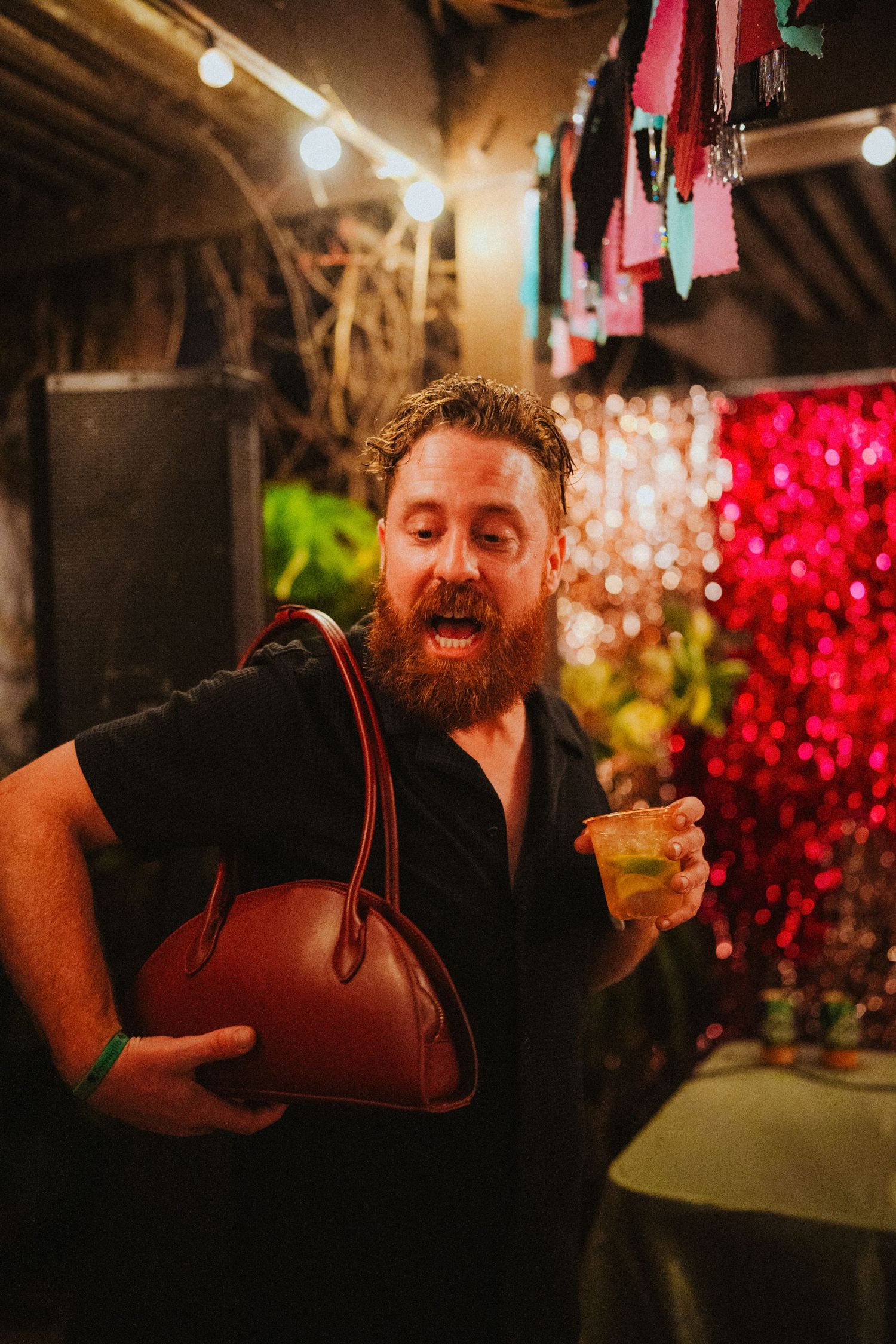 Bearded man in black shirt holds brown purse and drink at vibrant party with colorful decorations.