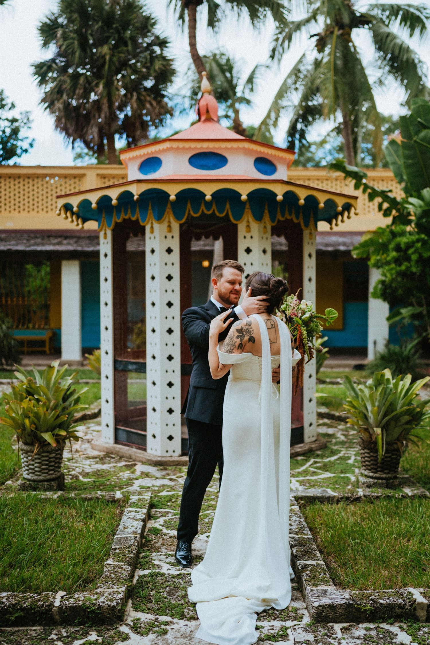 Bride and groom embrace in front of a colorful ornate gazebo surrounded by tropical palms.